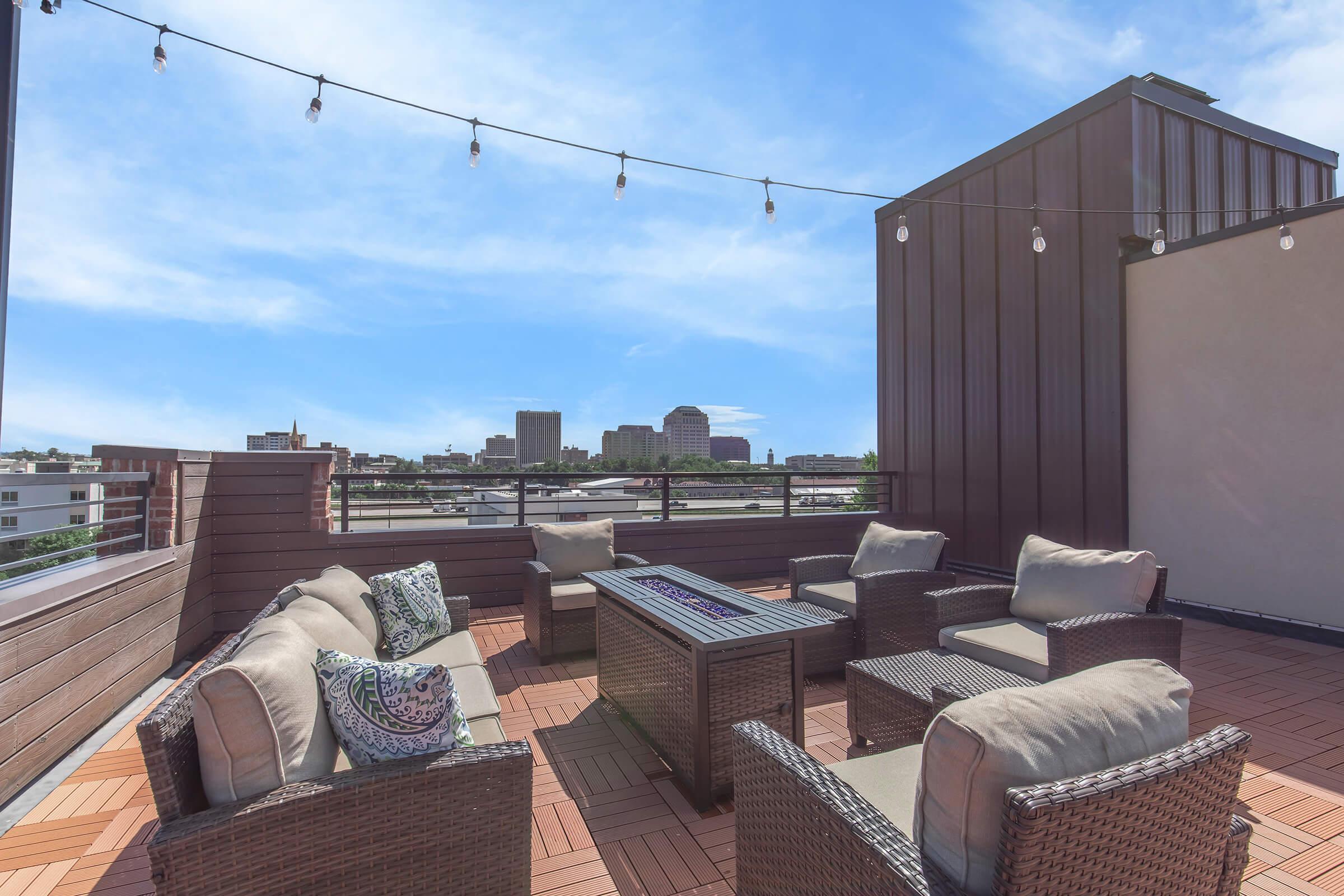A rooftop lounge area featuring comfortable seating with decorative pillows, a rectangular fire pit table, and string lights overhead. The backdrop includes a clear blue sky and a distant city skyline, creating a cozy and inviting atmosphere for relaxation or social gatherings.