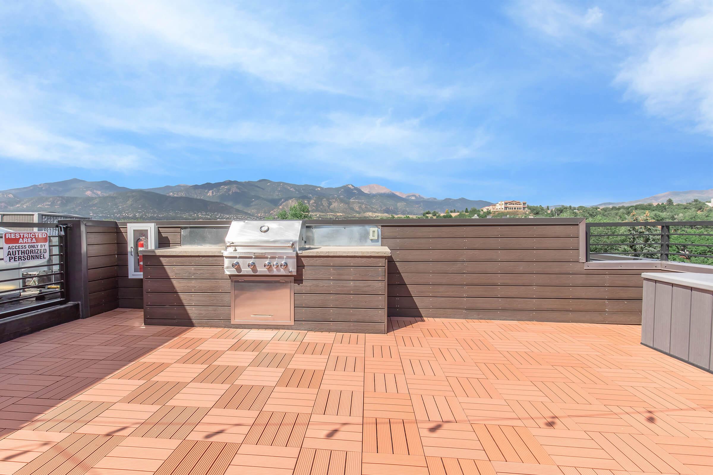 Outdoor rooftop patio featuring a built-in barbecue grill, surrounded by wooden fencing. The wooden deck has a herringbone pattern and offers stunning mountain views under a clear blue sky. A sign is posted nearby, and the area is well-lit, ideal for gatherings or relaxation.