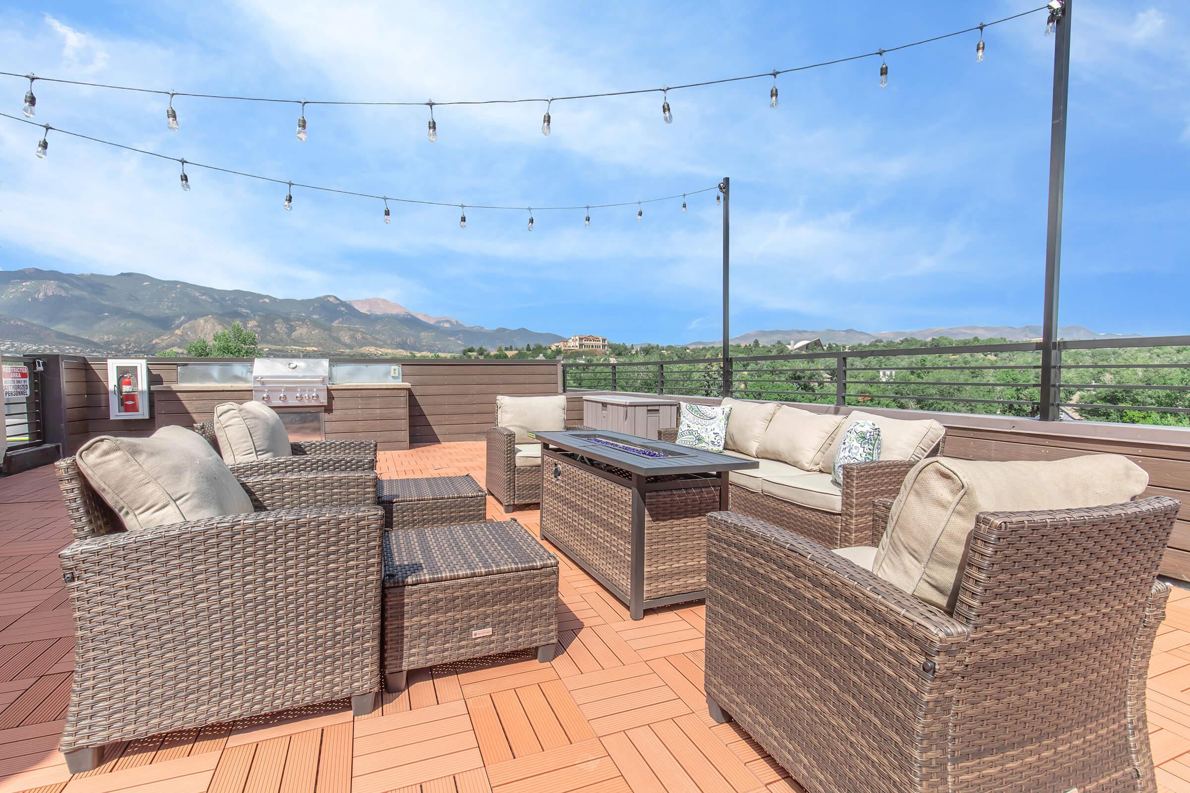 Outdoor patio area featuring comfortable wicker seating with cushions, a coffee table, and a grill in the background. The space is decorated with string lights above, set against a scenic view of mountains and a clear blue sky. Ideal for relaxation and gatherings.