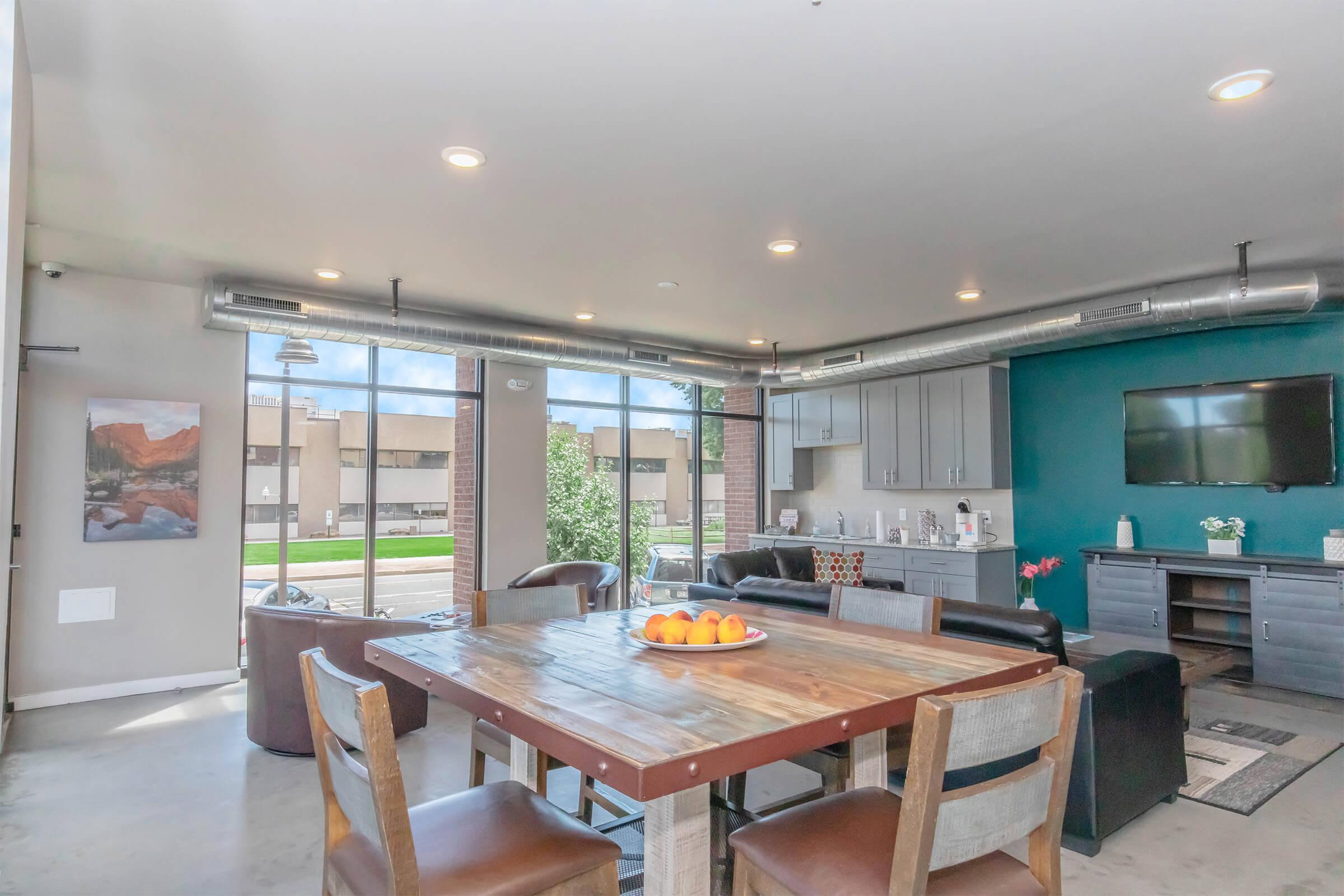 A modern communal living space featuring a wooden dining table with oranges, comfortable seating, and a large window providing natural light. A flat-screen TV is mounted on a teal wall, and there are cabinets and decor enhancing the inviting atmosphere.
