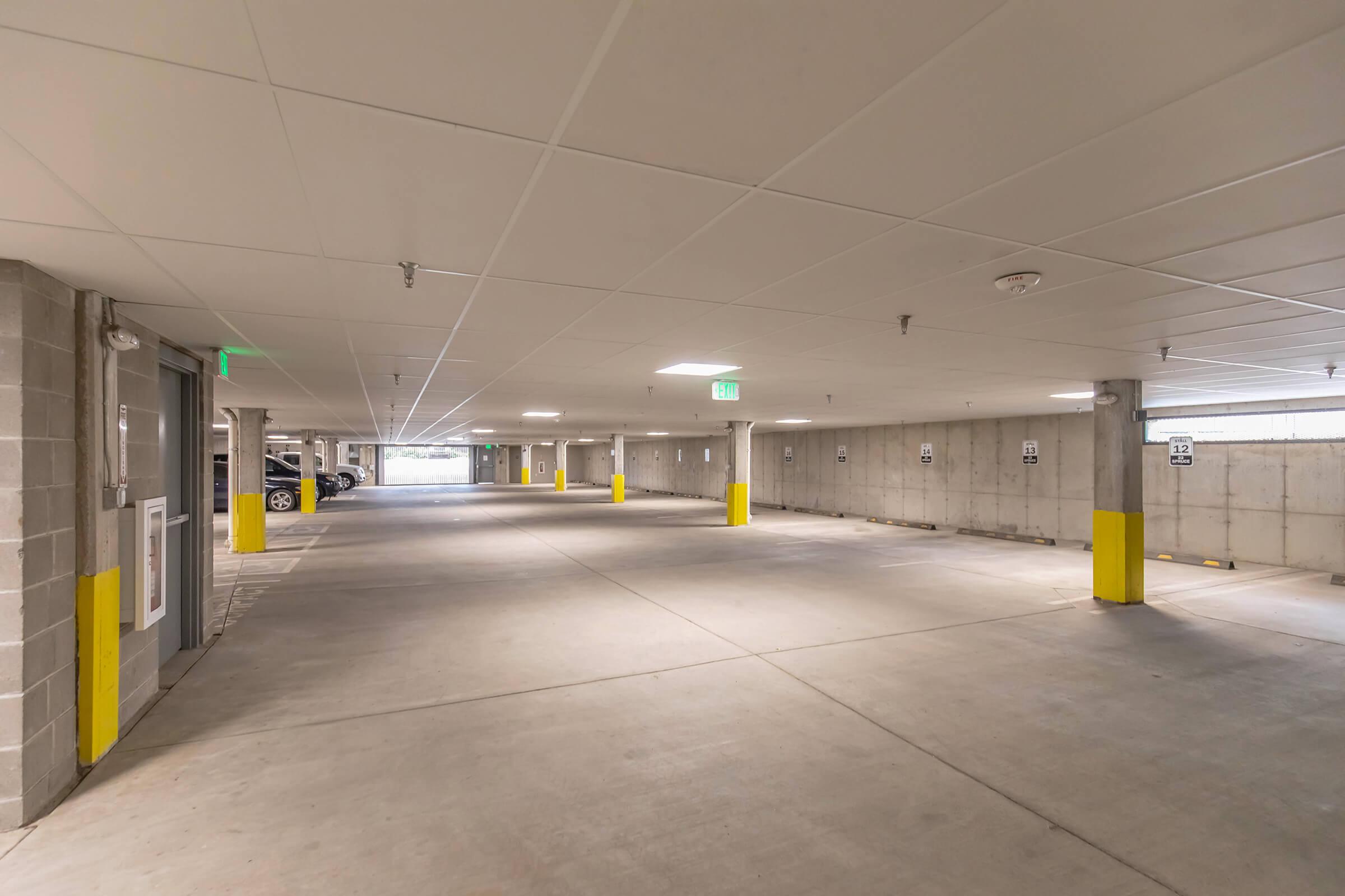 Empty underground parking garage with concrete walls and floors. Yellow columns mark parking spaces, and light fixtures illuminate the area. A few parked cars are visible in the distance. The space appears clean and well-maintained, with signs indicating parking regulations.