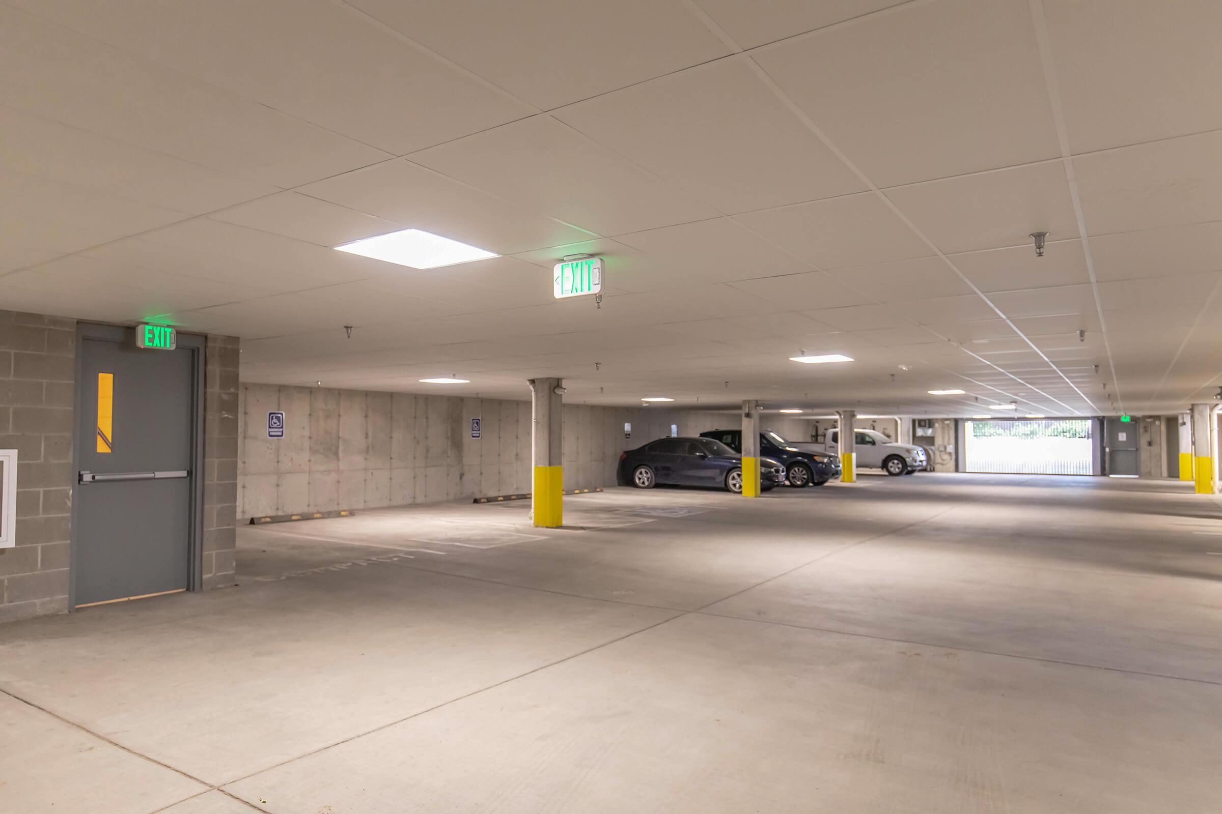A spacious, well-lit indoor parking garage with concrete walls and columns. There are yellow safety posts near parked cars, and exit signs illuminated on the ceiling. The area appears clean and organized, with a couple of cars parked in designated spots.