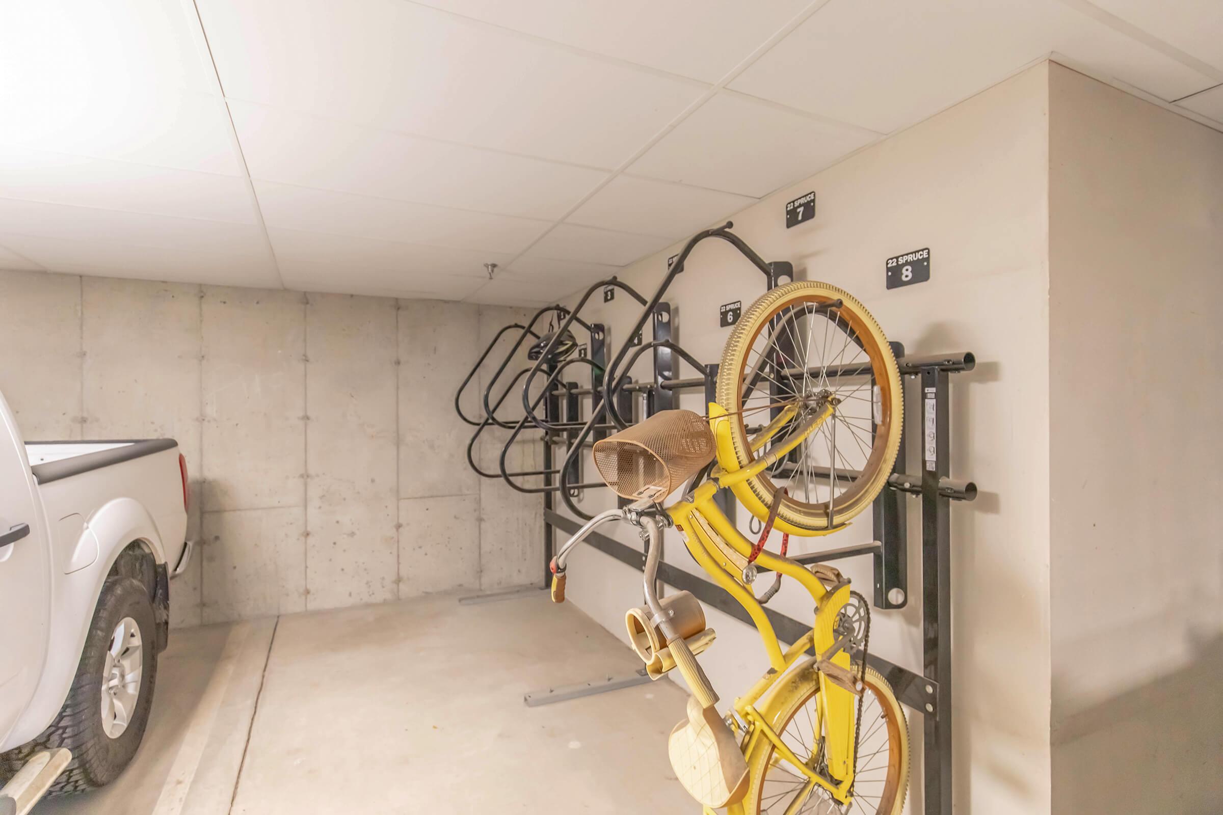 A bright underground garage featuring concrete walls and a clean floor. On the left, a white vehicle is parked. The right wall displays several bike racks with a yellow bicycle hanging on one of them, showcasing its unique design. The space is organized and well-lit, with identification numbers visible on the walls.
