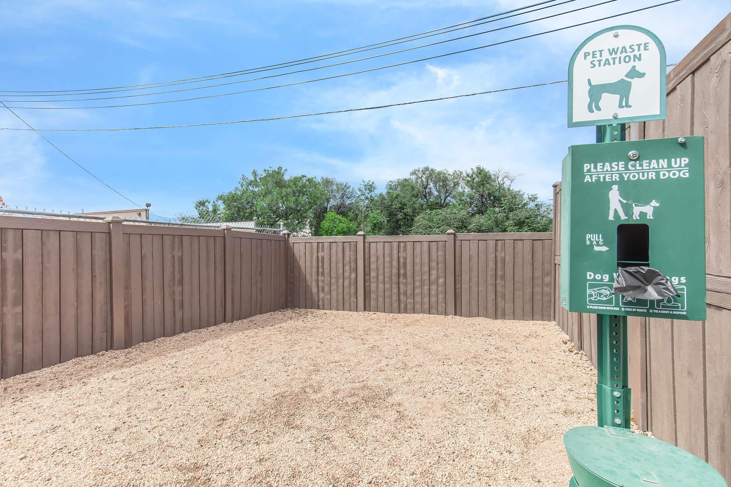 Pet waste station in a fenced area with a sign encouraging dog owners to clean up after their pets. The station features a bag dispenser and a trash receptacle, surrounded by a gravel surface and wooden fencing under a clear blue sky.