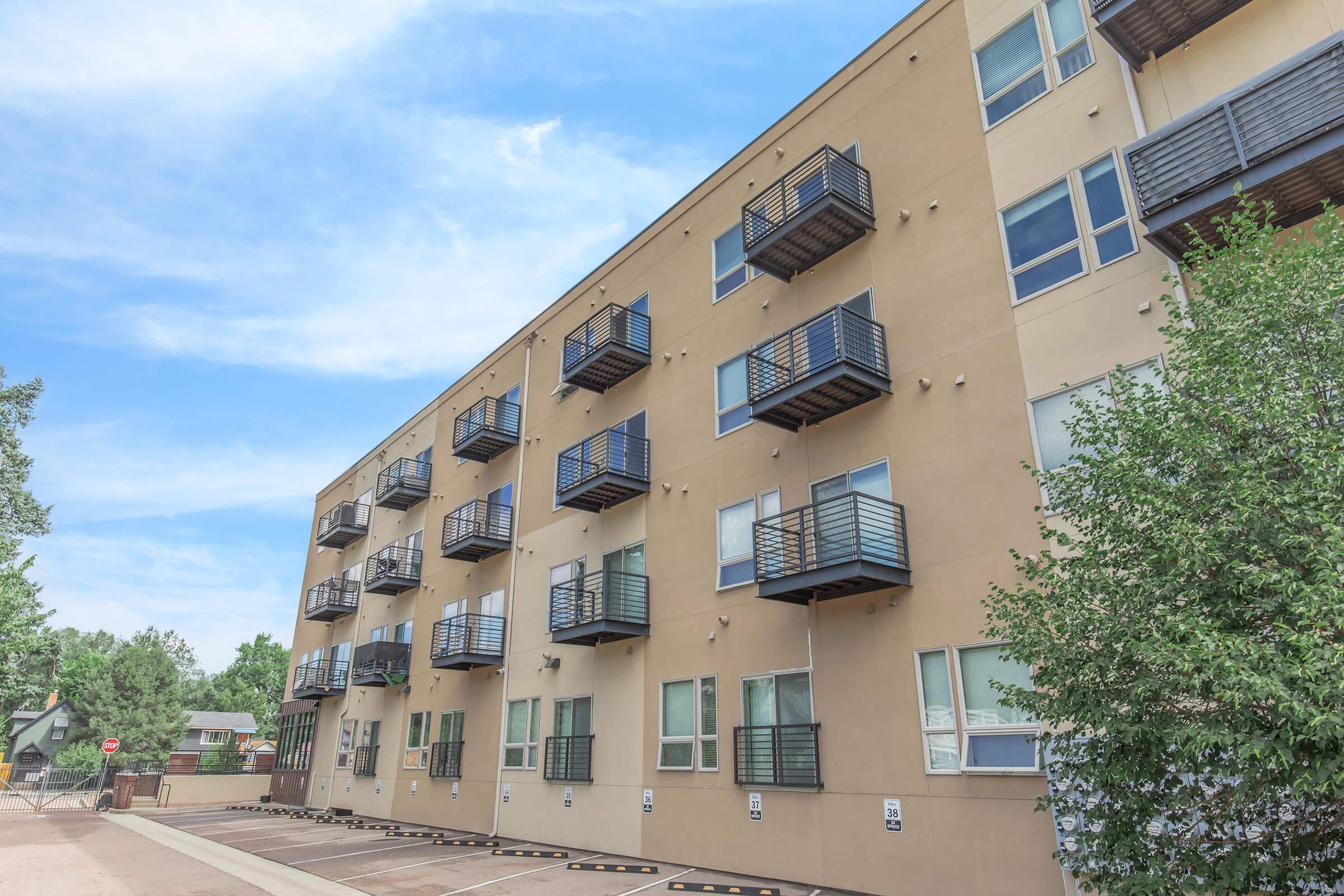 Exterior view of a modern apartment building with several balconies. The facade is beige with large windows. There are trees and a paved area in front, showcasing a clear blue sky in the background. Parking spaces are visible along the building's side.
