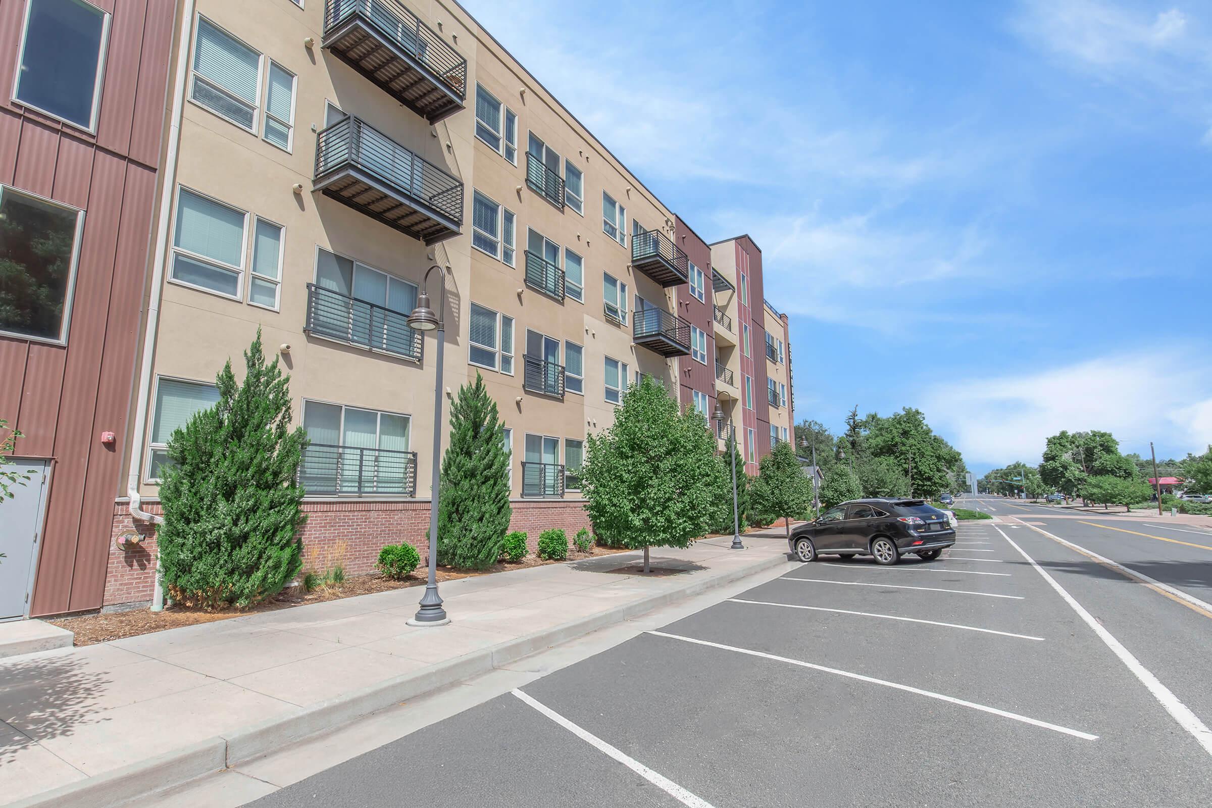 A modern apartment building with multiple floors featuring balconies, surrounded by neatly landscaped greenery. A black car is parked alongside a wide street with empty parking spaces, under a blue sky with a few clouds. The scene conveys a peaceful urban setting.