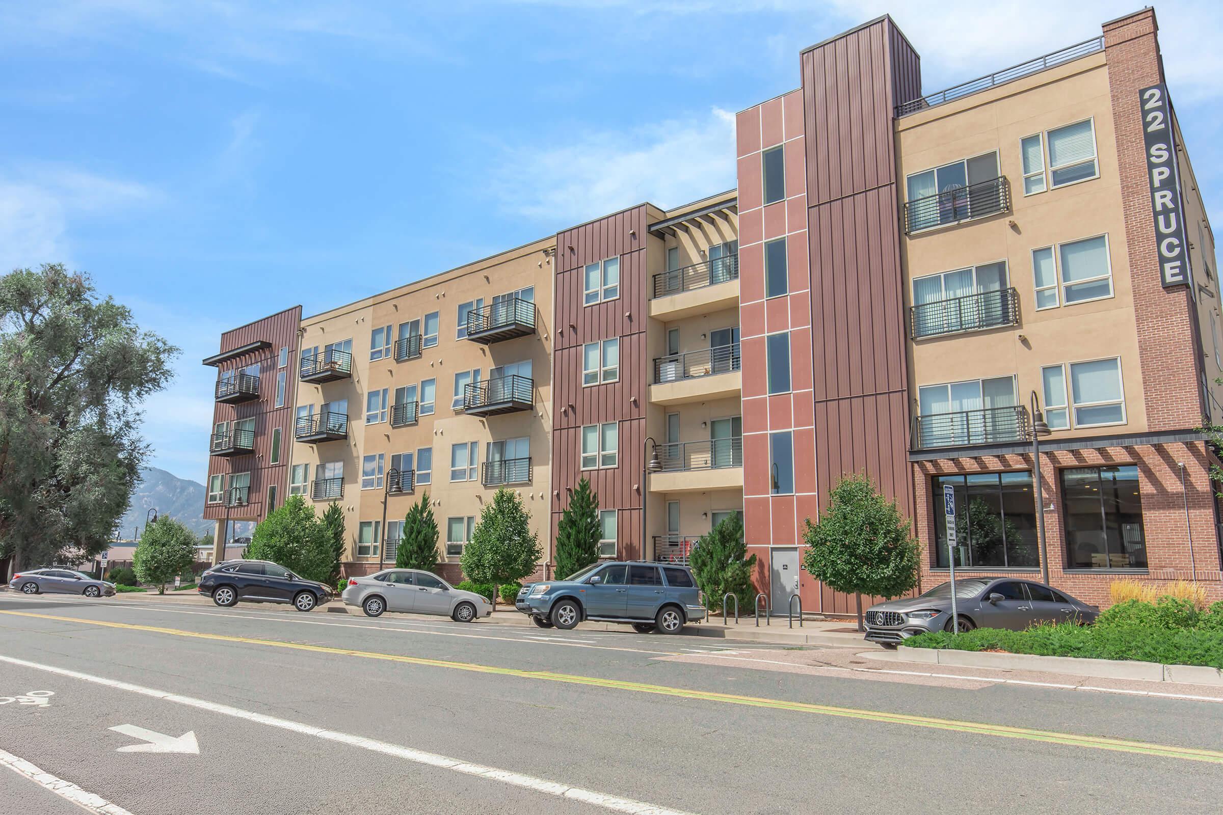 A modern apartment building with a mix of beige and brown exterior. It has multiple balconies visible on the upper floors, lined with small trees in front. Several parked cars are on the street in front of the building, which features a sign that reads "22 Spruce." Bright blue sky is in the background.