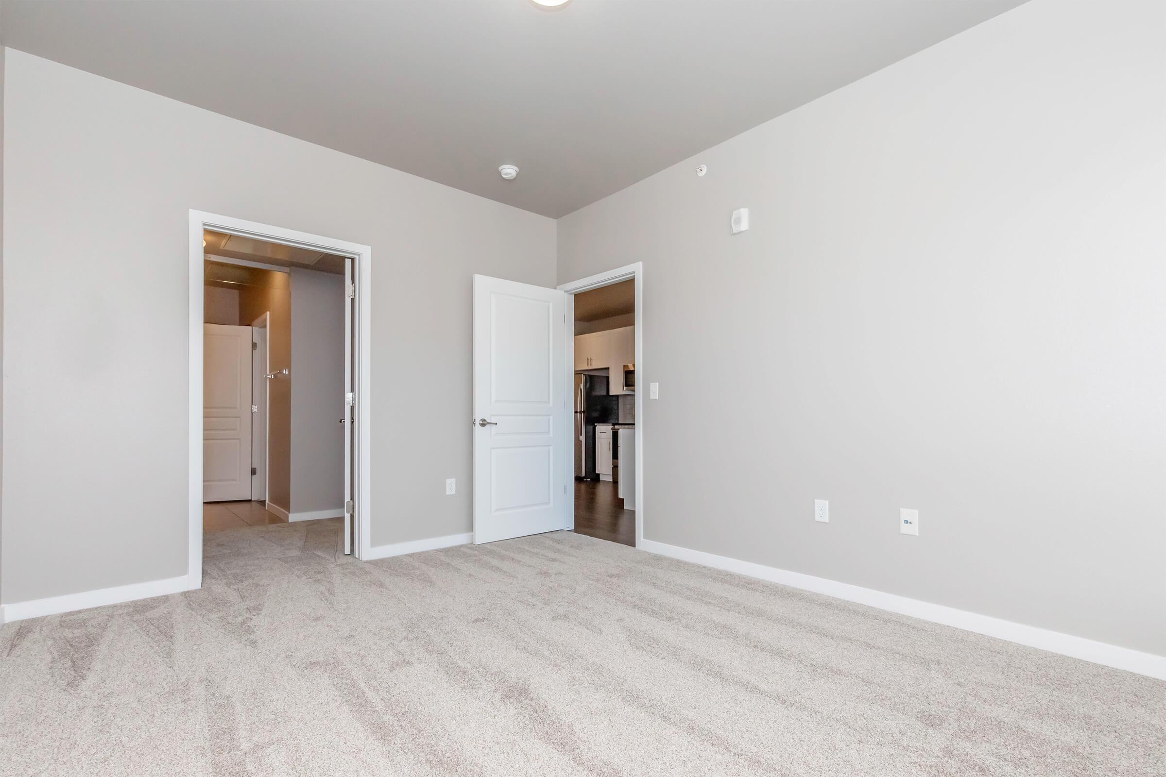 A spacious, empty room with light gray walls and carpeted flooring. There are two doors: one leading to a bathroom and another opening to a kitchen area. Natural light enters through the doorways, creating a bright atmosphere. The room is ready for furnishings and personal decor.