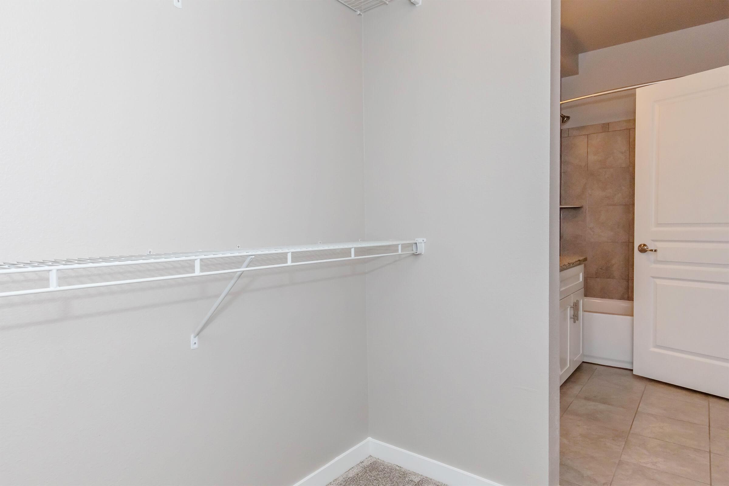 Empty closet space featuring a white wire shelf on a light gray wall, with light-colored tile flooring. A bathroom is visible in the background, partially obscured. The overall aesthetic is clean and modern.