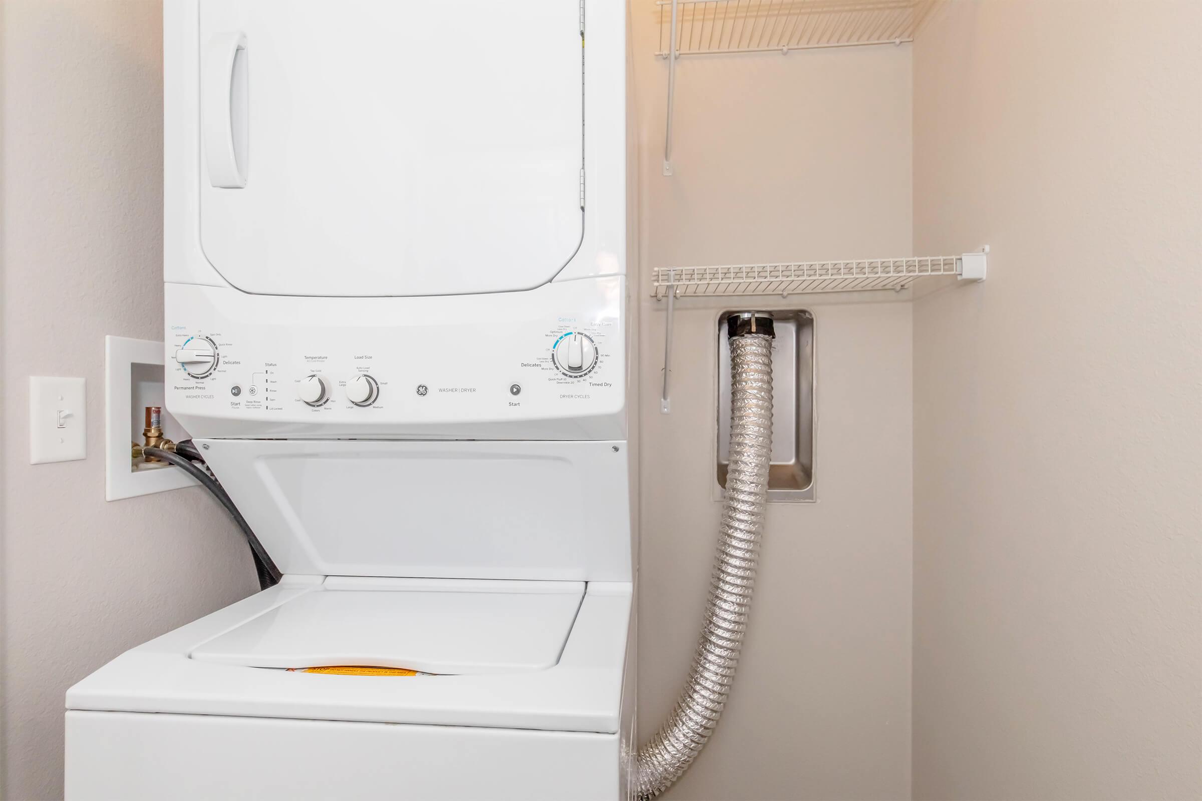 A stacked washer and dryer unit in a laundry space, with controls visible on the front of the washer. The dryer vent is connected to a wall and there's a shelf above for storage. The walls are painted a light color, creating a clean and tidy appearance.
