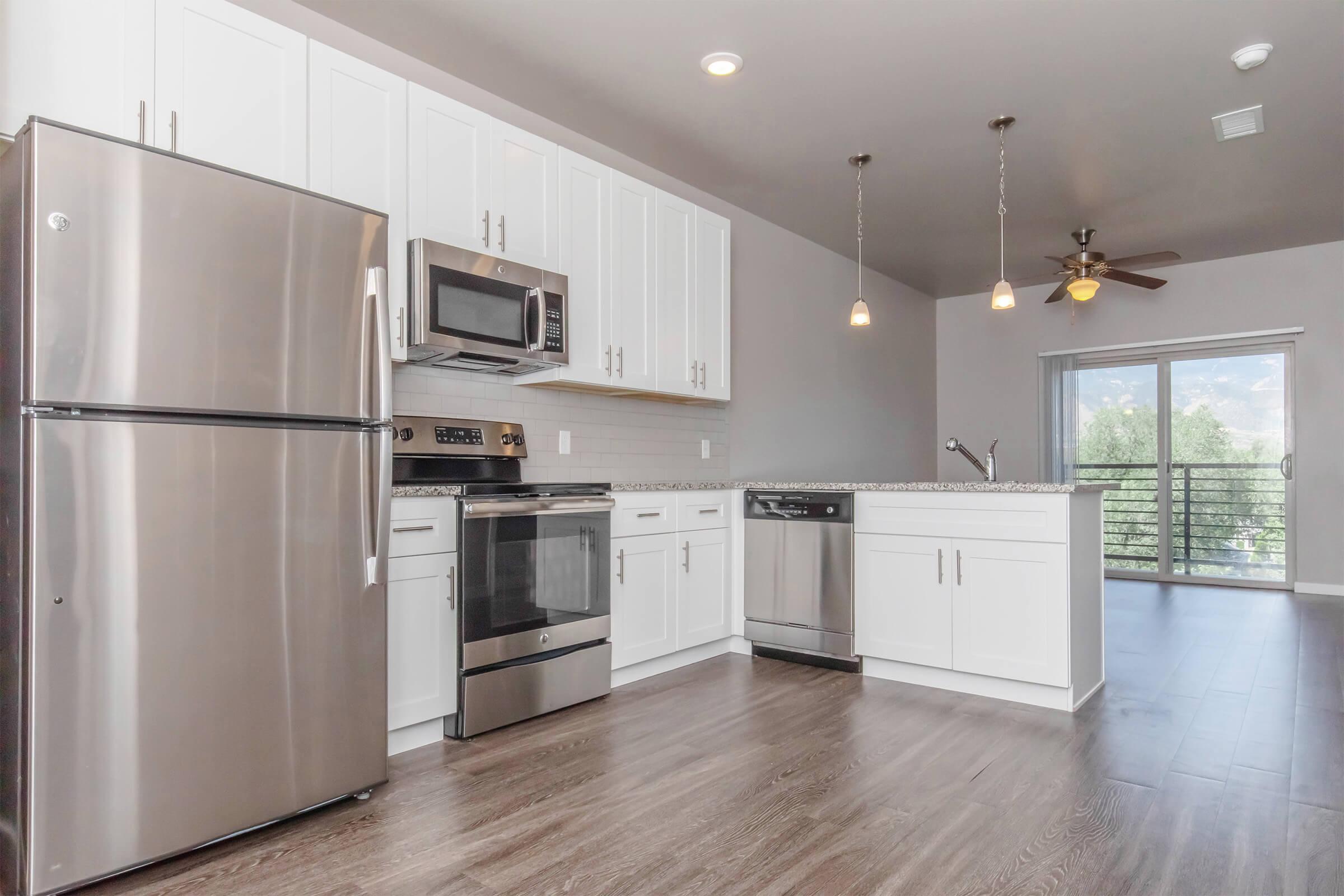 A modern kitchen featuring stainless steel appliances, including a refrigerator, oven, microwave, and dishwasher. The cabinets are white, and there is a granite countertop. Soft pendant lights hang above the counter, and a sliding glass door leads to a balcony with a view. The flooring is a dark wood finish.