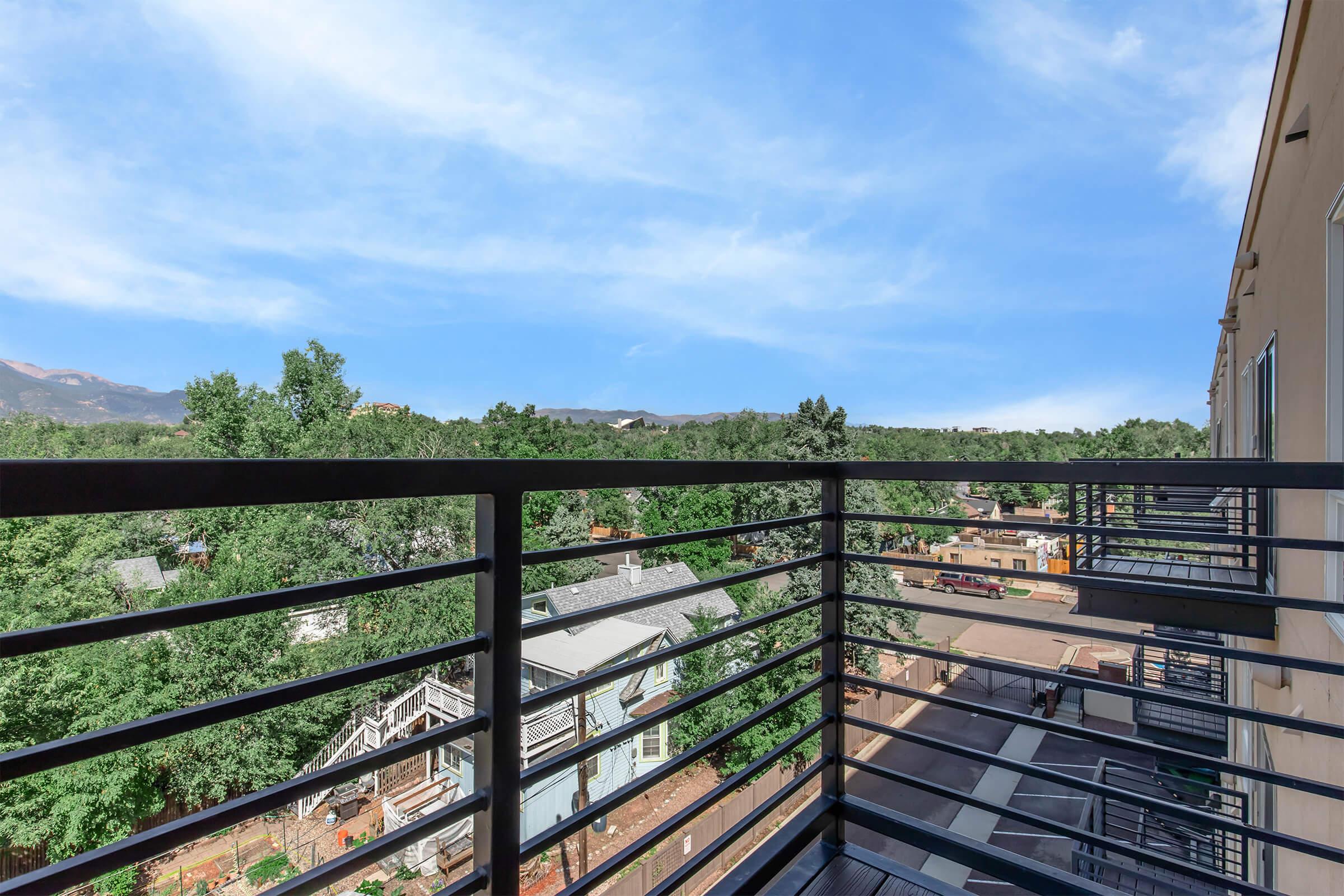 View from a balcony overlooking a scenic landscape with trees and distant mountains. The foreground features a black metal railing, while the background shows a mix of greenery and residential buildings. The sky is clear with a few clouds, creating a peaceful atmosphere.