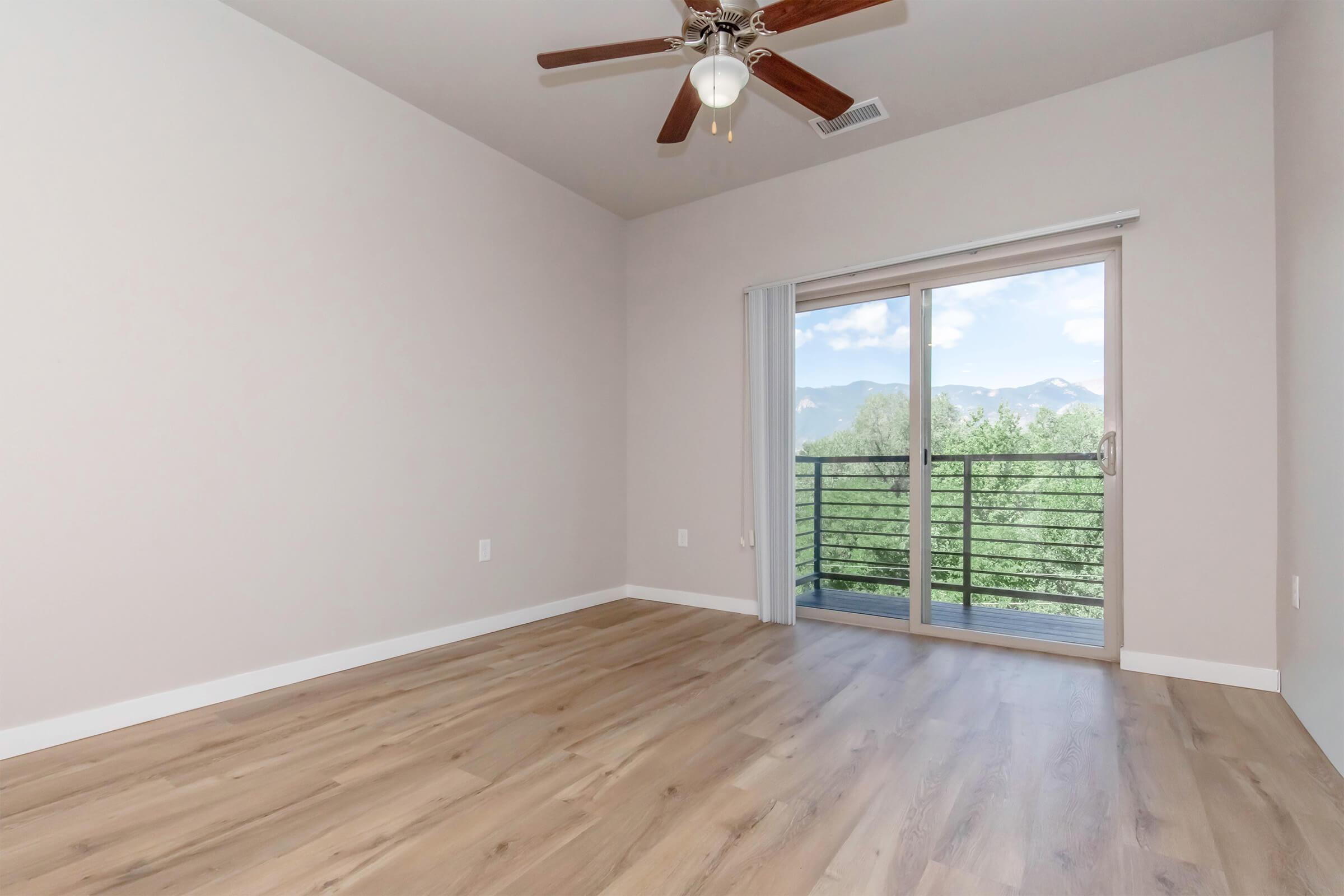 A spacious, empty room with light-colored walls and a ceiling fan. Large sliding glass doors lead to a balcony overlooking trees and mountains. The floor is made of light wood, creating a bright and airy atmosphere. Natural light floods the space from the balcony.