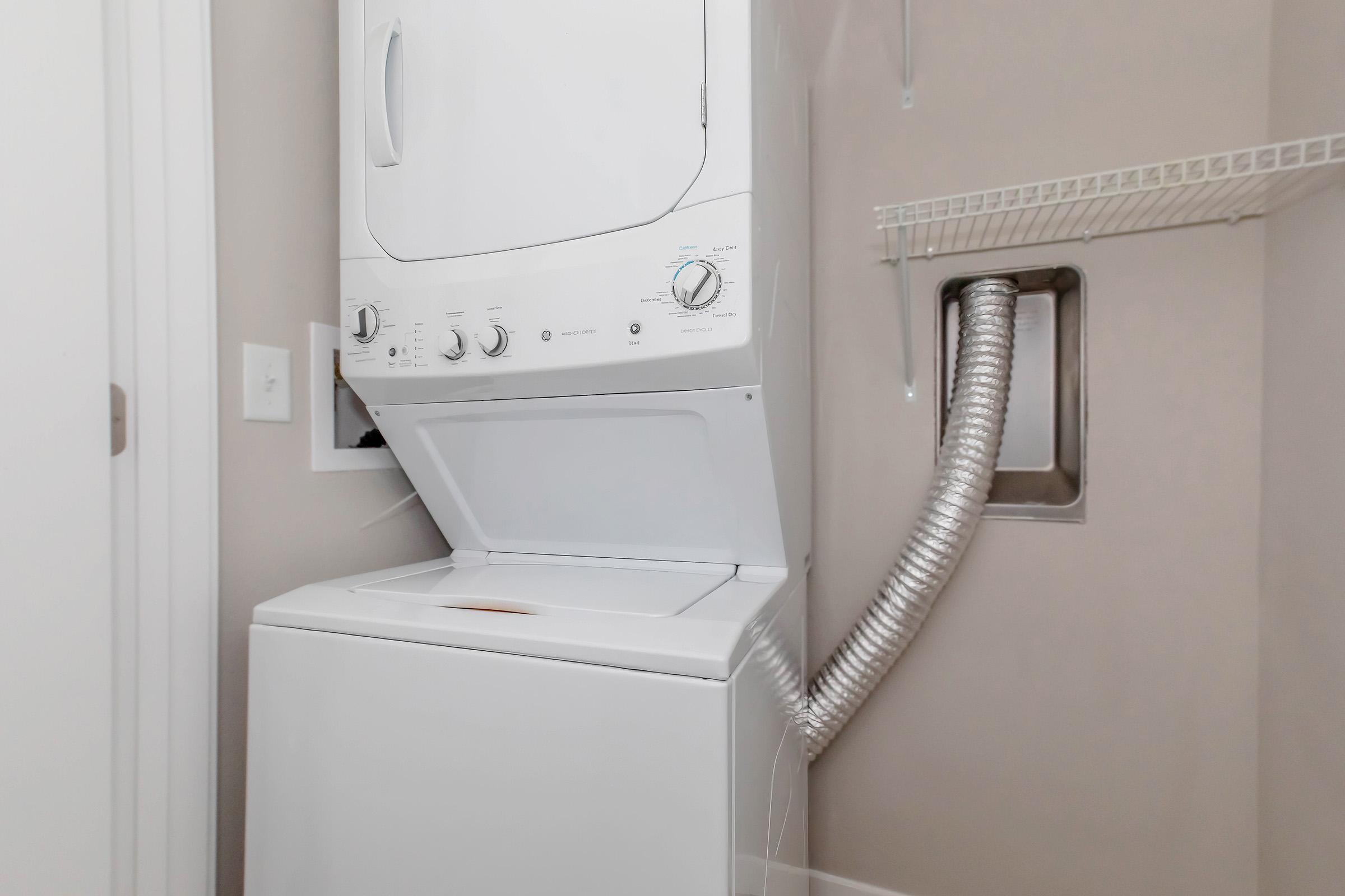 A stacked washer and dryer unit in a small laundry space, with a vent hose attached to the wall. The washer is on the bottom, and the dryer is on top, both featuring control dials and a clean, modern design. The background includes a light-colored wall and a shelving unit above.