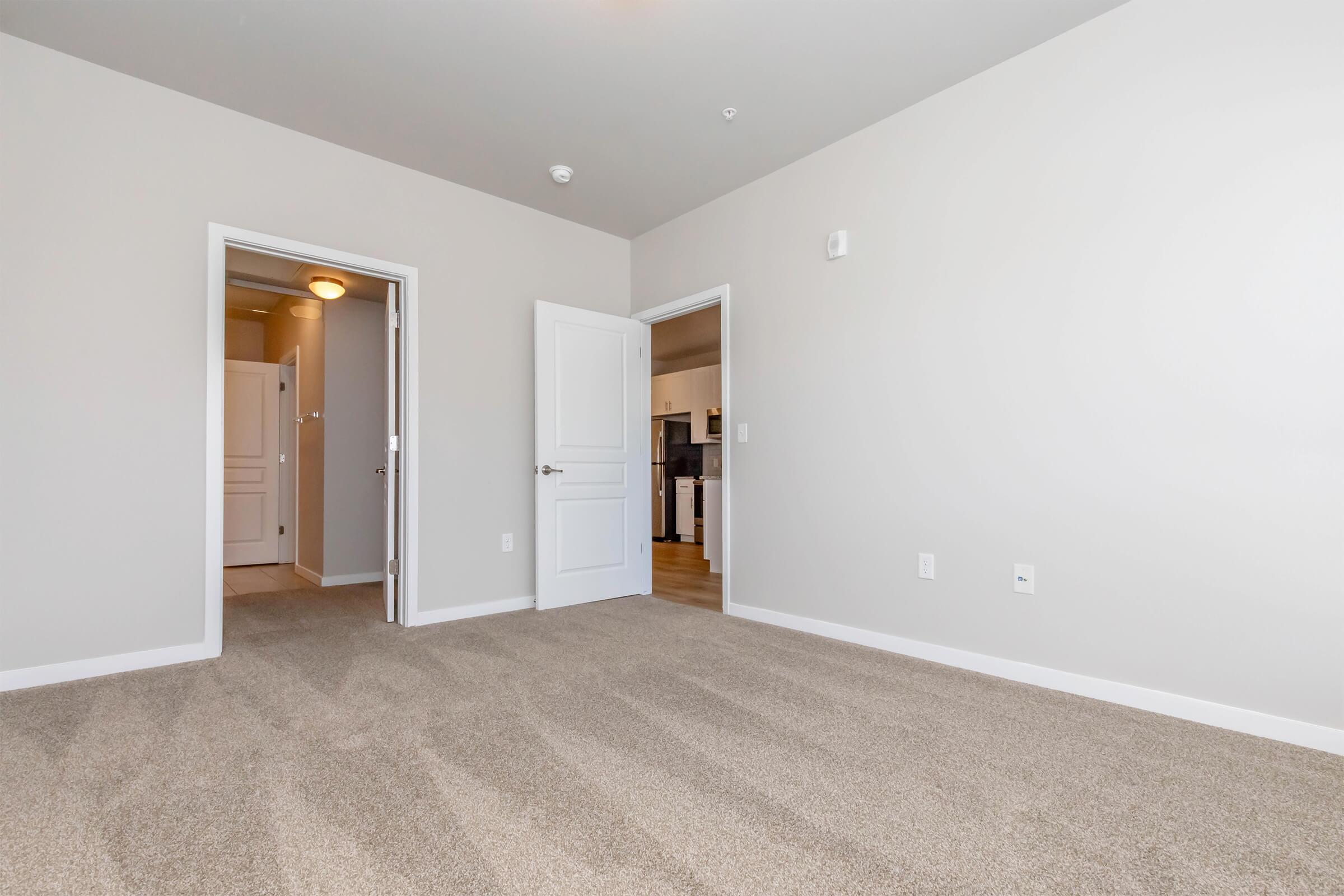 Empty room with light beige walls and carpeted floor. Two doorways are visible—one leading to a hallway and the other to a kitchen area. The space is well-lit with natural light, creating a spacious and inviting atmosphere. The overall aesthetic is modern and minimalist.
