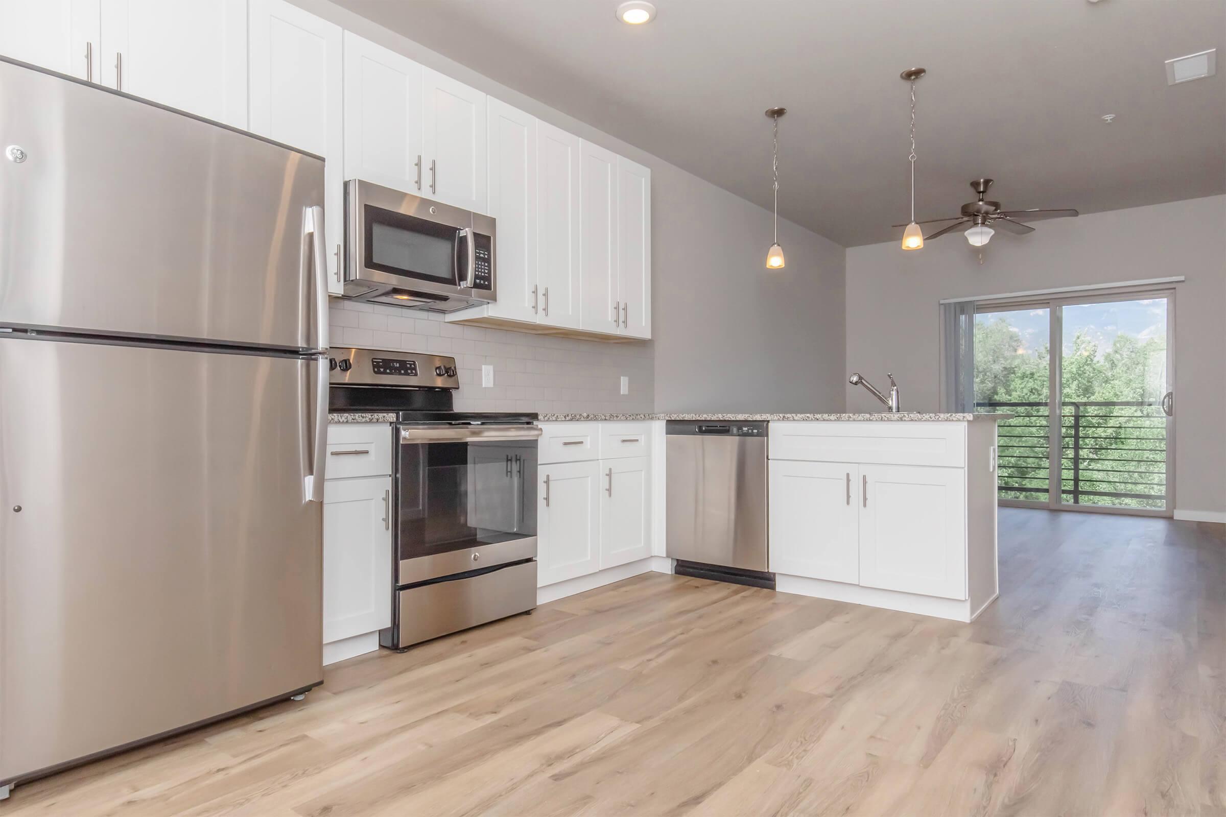 Modern kitchen featuring stainless steel appliances, including a fridge, oven, and microwave. White cabinets and a granite countertop complement the design. The open layout leads to a living area with a view of an outdoor balcony through sliding glass doors. Light wooden flooring throughout.