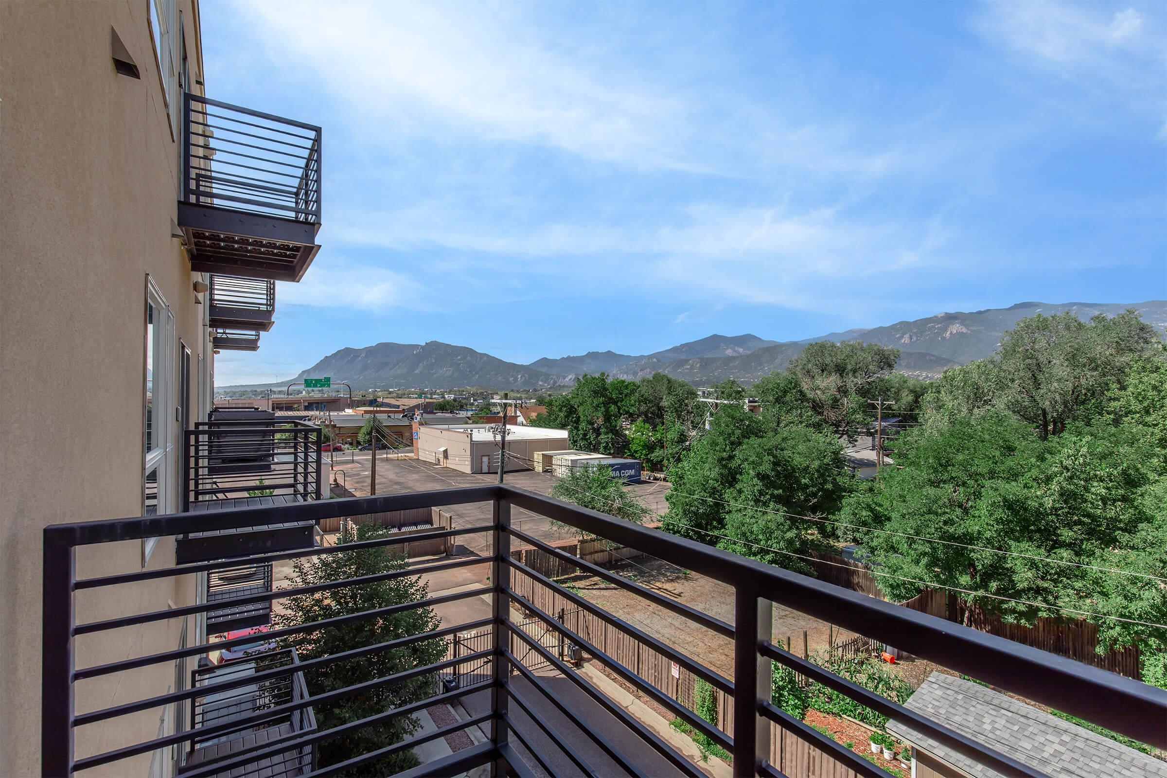 A scenic view from a balcony overlooking a tree-lined street with mountains in the background. The image captures buildings and greenery, showcasing a bright blue sky with light clouds. The balcony's metal railing adds depth to the composition, emphasizing the outdoor space.