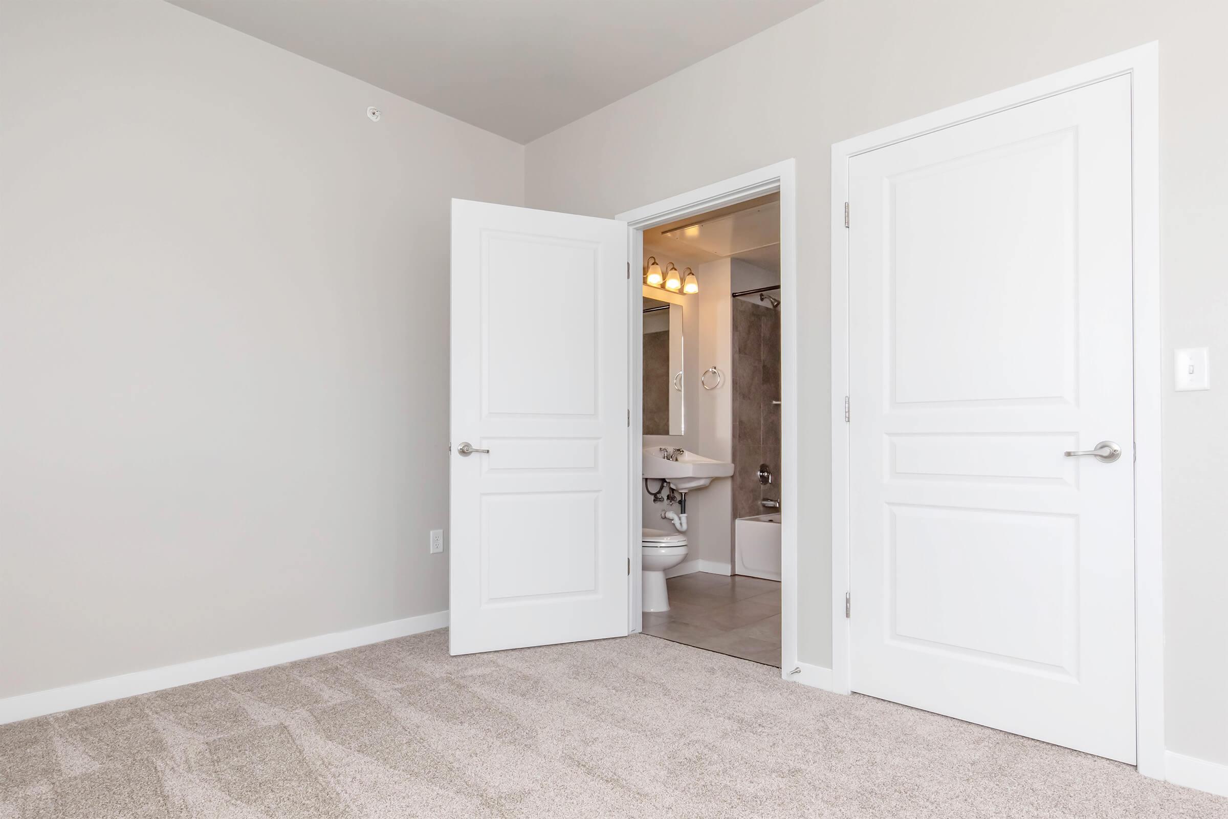 A bright, neutral-colored room with freshly painted walls and plush carpet. A partially open door leads to a bathroom featuring modern fixtures, including a sink and bathtub. The overall atmosphere is clean and inviting, suitable for a living space.