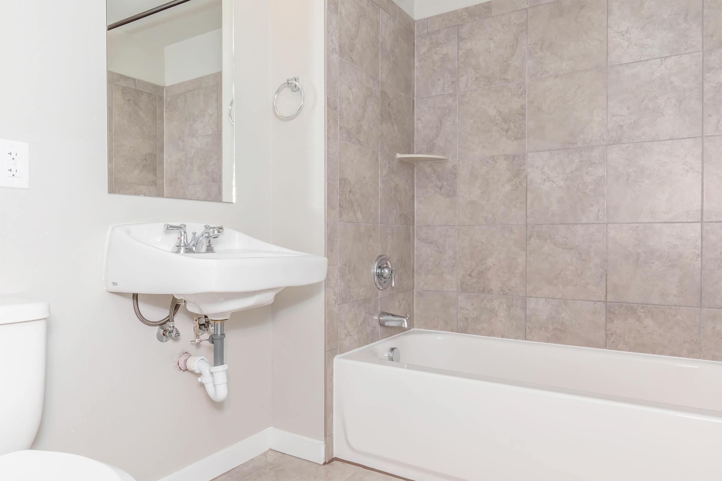 A small bathroom featuring a white bathtub, a wall-mounted sink with a faucet and plumbing visible beneath, and a modern mirror. The walls are tiled in a light gray pattern, and there is a chrome towel holder. The floor is light-colored, and the overall design is clean and minimalistic.