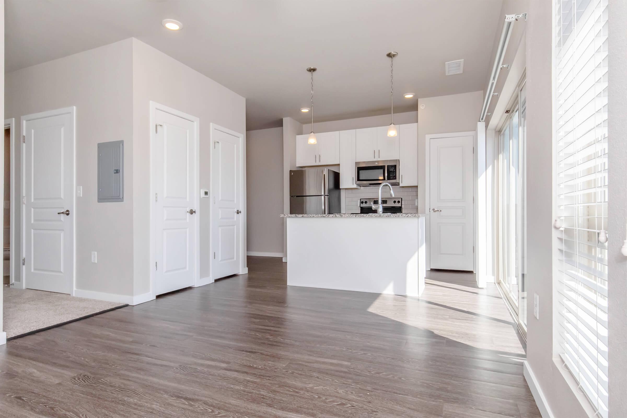 A spacious, modern kitchen area with an open floor plan. It features light-colored walls, wooden flooring, and a central island with a granite countertop. There are stainless steel appliances, including a refrigerator and microwave, and multiple doors leading to adjacent rooms. Large windows allow natural light to fill the space.