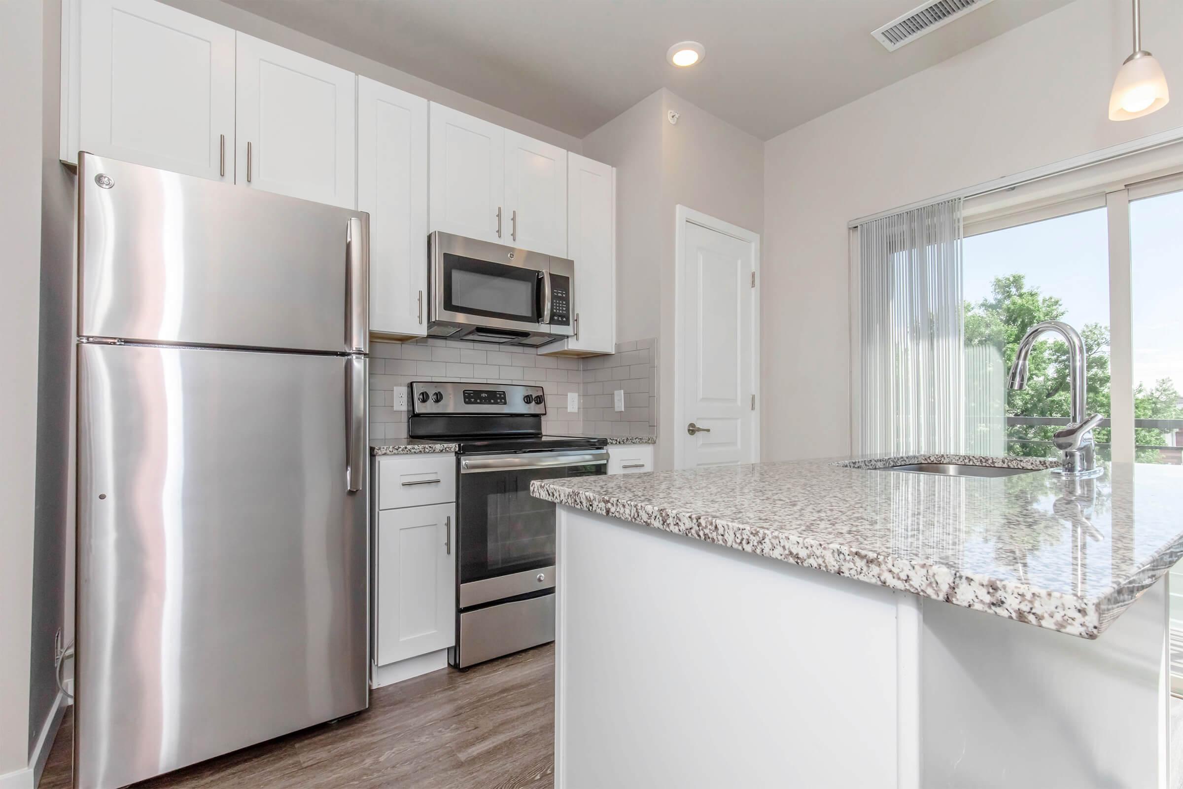 Modern kitchen featuring stainless steel appliances, including a refrigerator, microwave, and stove. The kitchen has white cabinets, a granite countertop, and a spacious island. Large window allows natural light, giving a bright and airy feel to the space.
