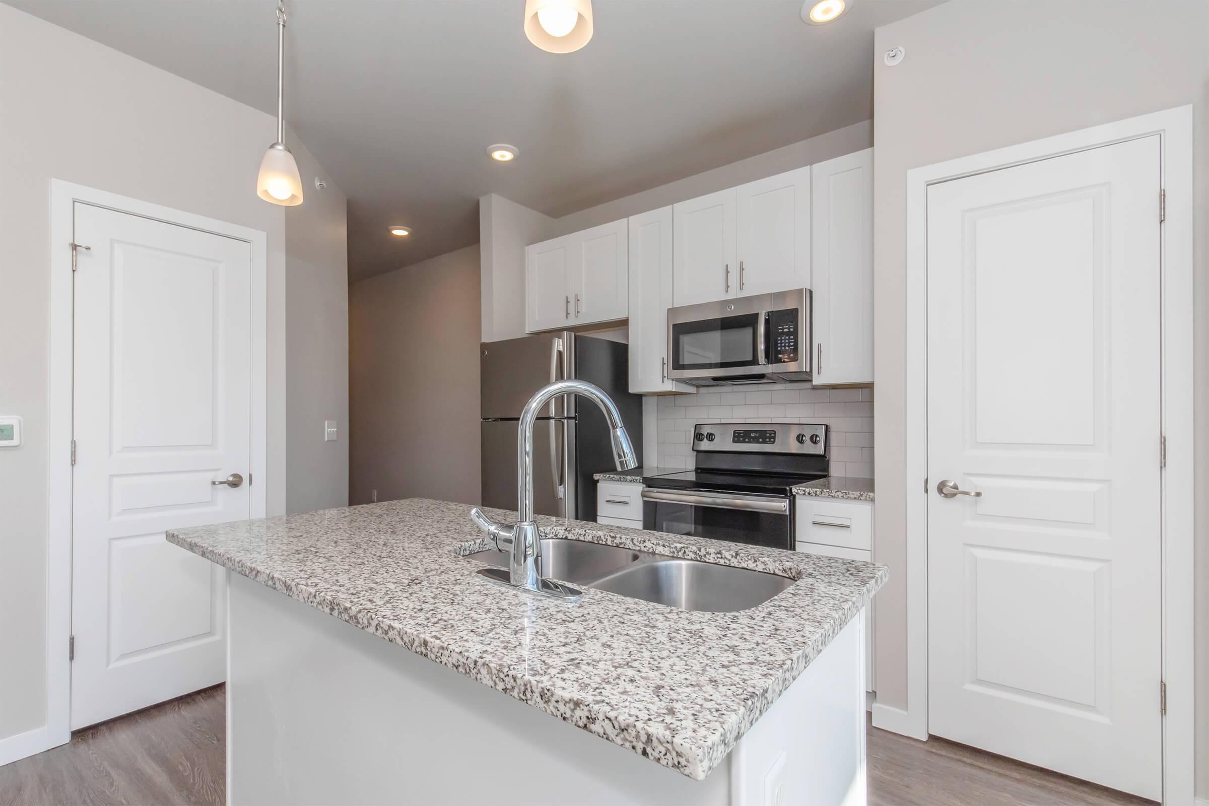 A modern kitchen featuring white cabinetry, stainless steel appliances, and a granite countertop. The kitchen includes a sink on an island, pendant lighting, and an open layout with a light color scheme, creating a bright and inviting atmosphere.