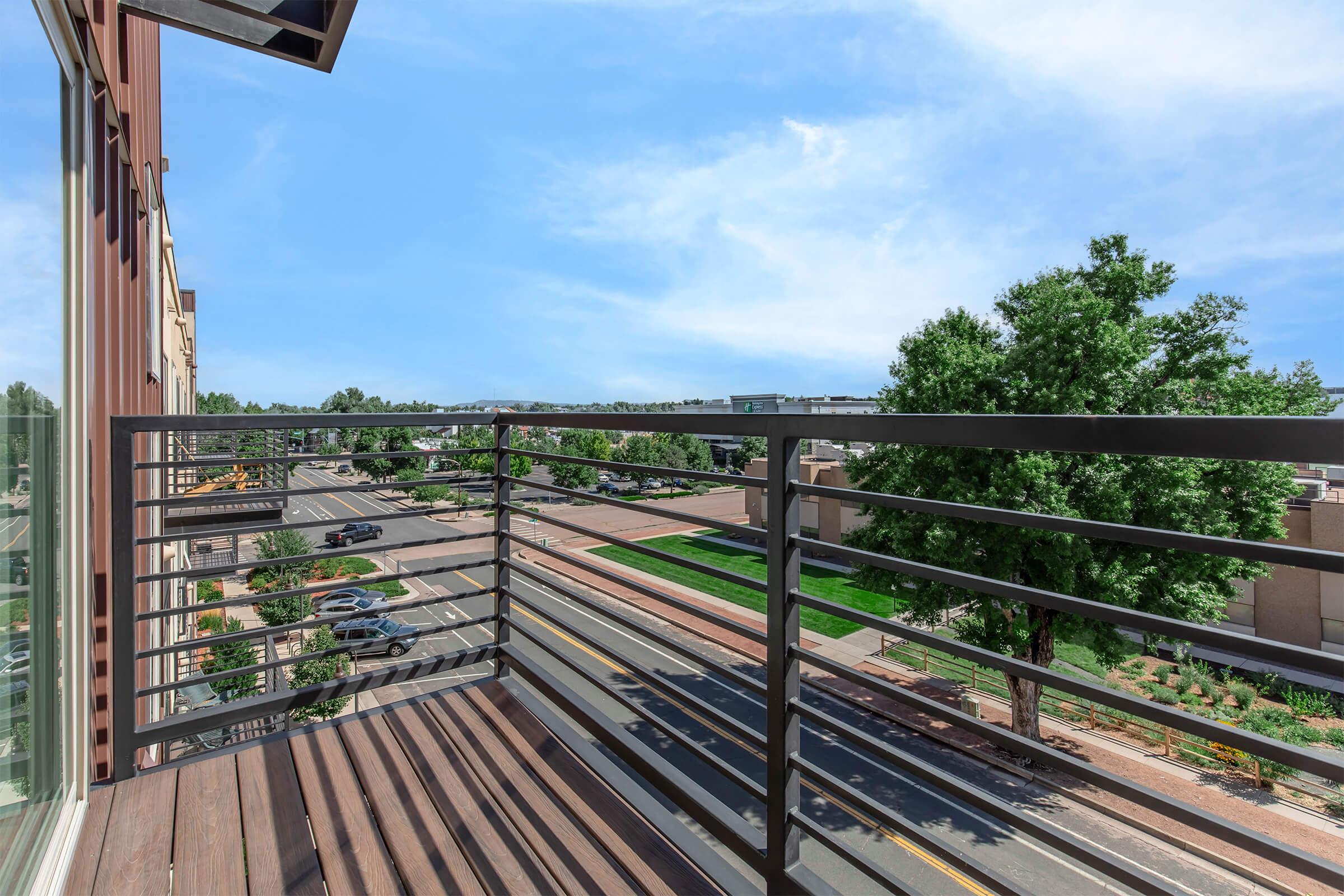 A view from a balcony overlooking a tree-lined street. The railing features a modern design, with a clear blue sky in the background. The scene includes parked cars along the road and green landscaping visible in the foreground.