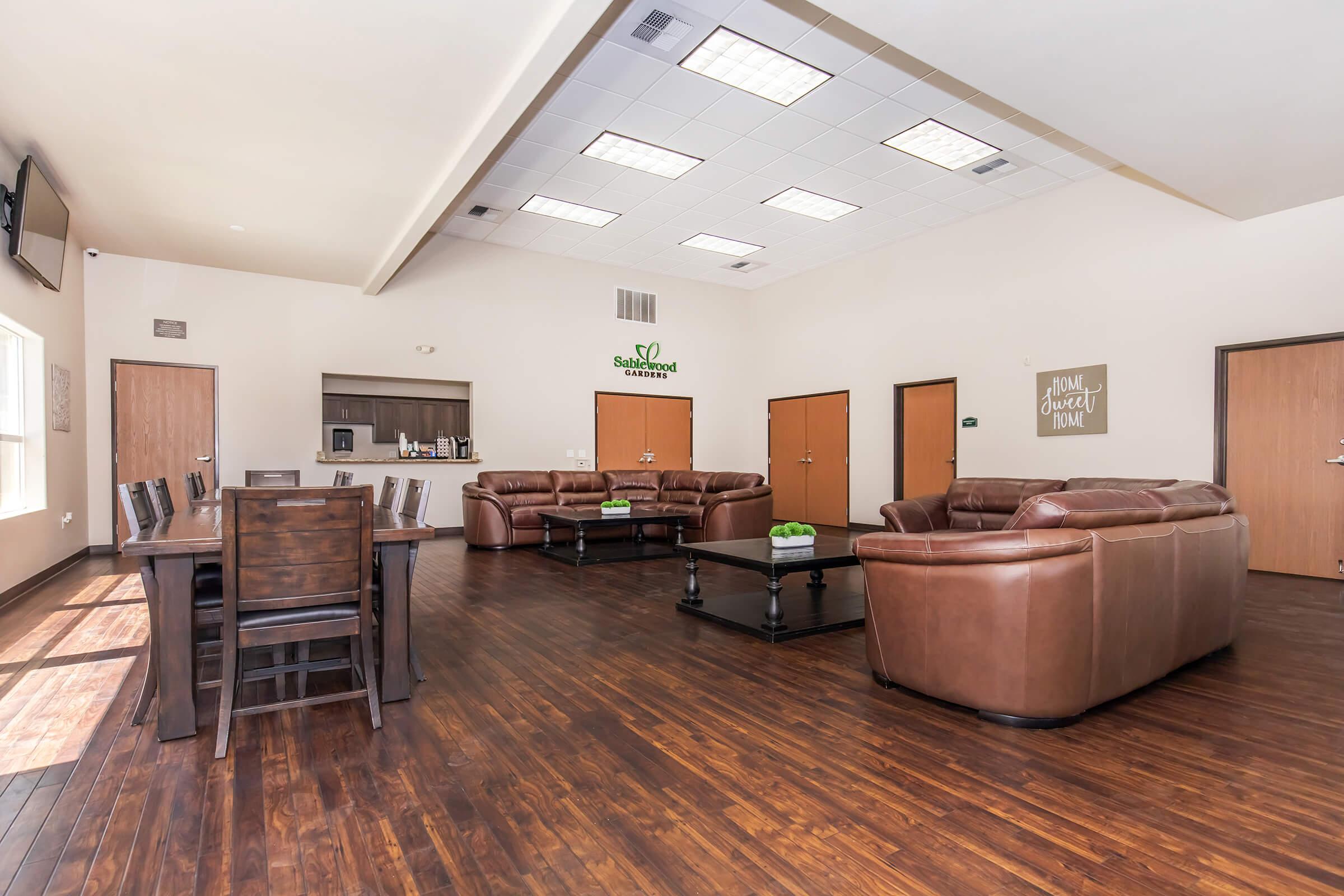 Spacious communal lounge with hardwood flooring, featuring two brown leather sofas, a coffee table, and a dining area with a large wooden table and chairs. There is a kitchenette visible in the background, along with neutral-colored walls and minimal decor. Natural light brightens the room through large windows.