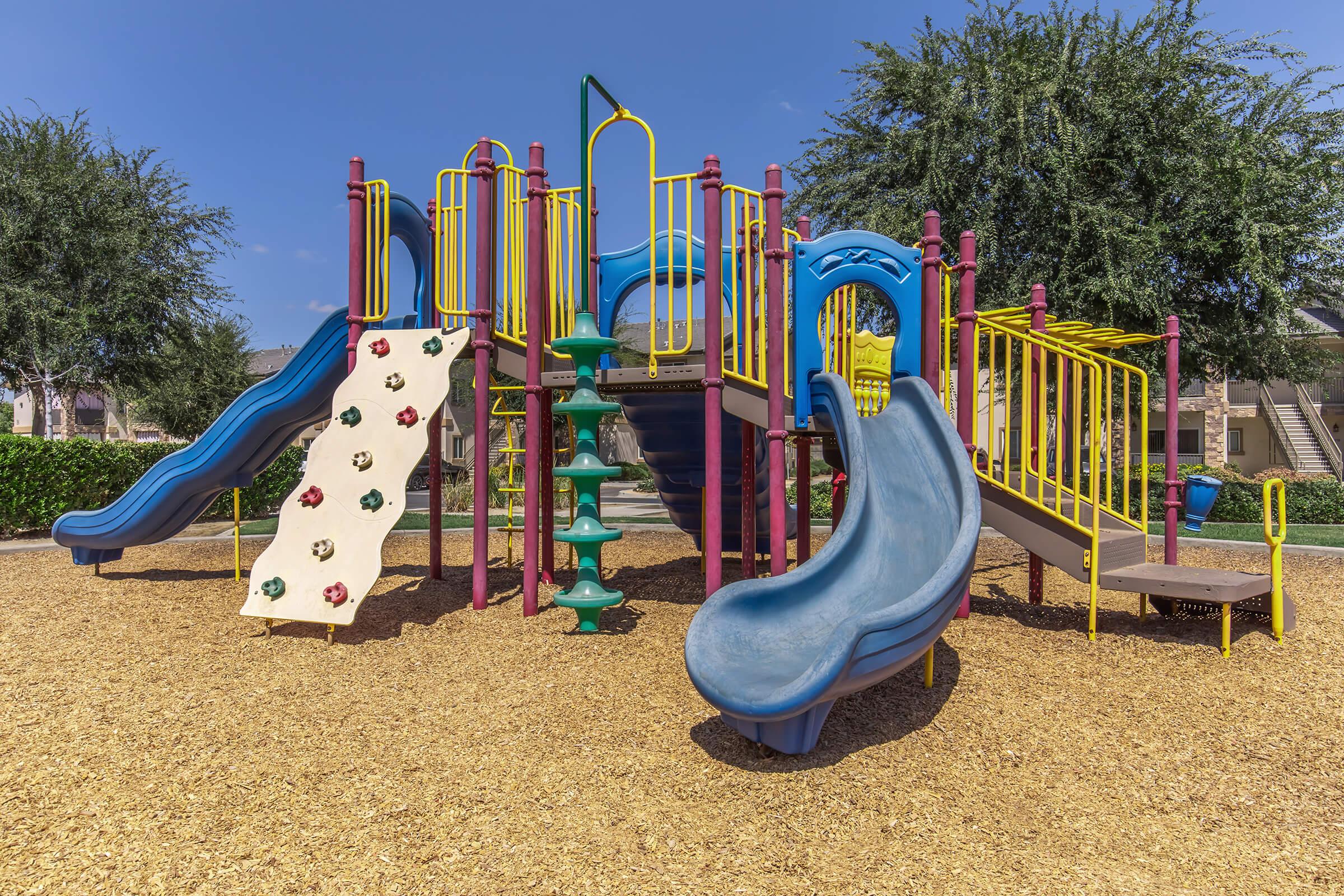 Colorful playground equipment featuring multiple slides, climbing structures, and a rock wall, set against a clear blue sky. Surrounding the playground are trees and a carpet of wood chips, creating a safe play area for children.