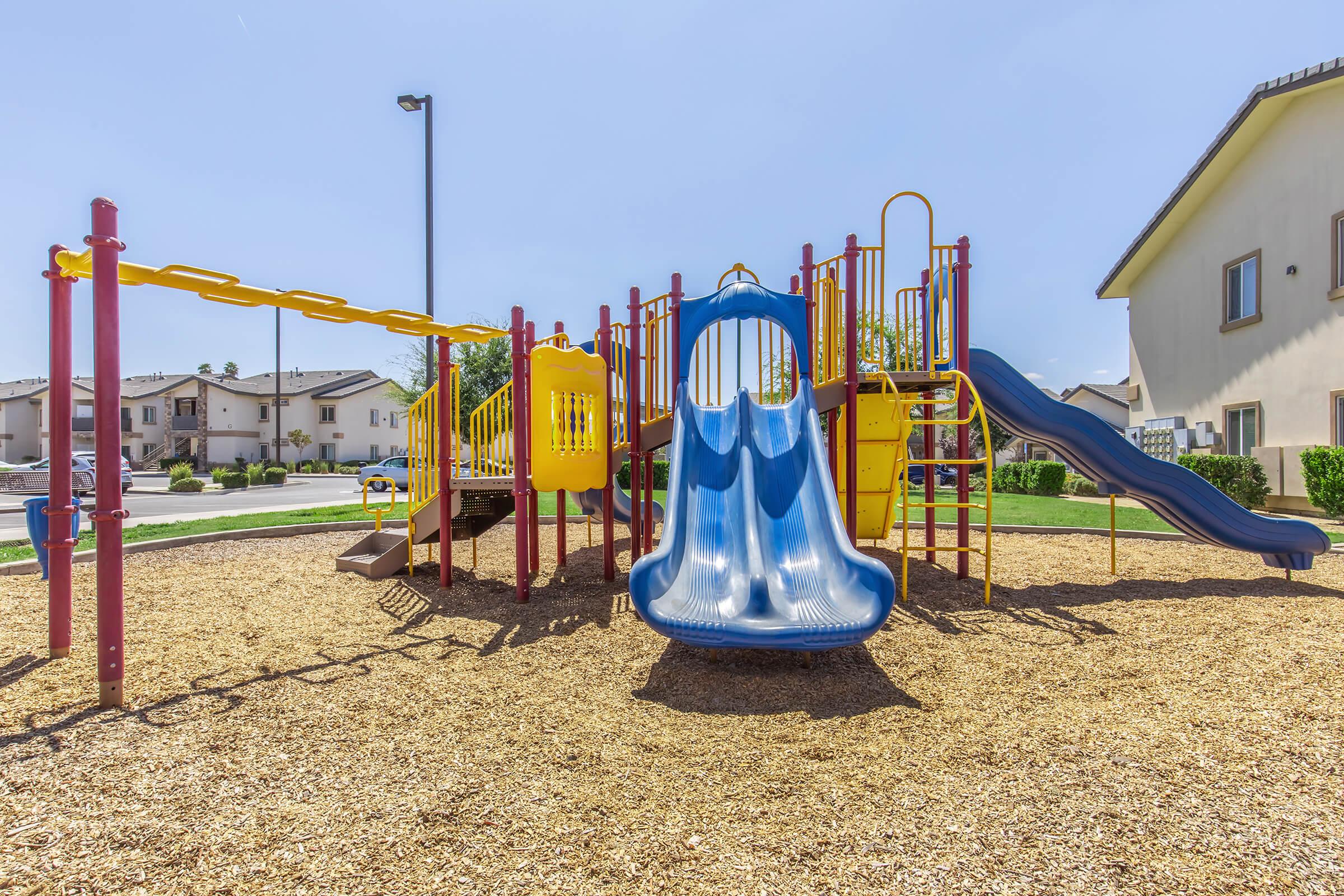 A colorful playground featuring a blue slide, climbing structures, swings, and safety railings, surrounded by a sandy surface. In the background, residential buildings are visible under a clear blue sky. The area is well-maintained and designed for children's play.