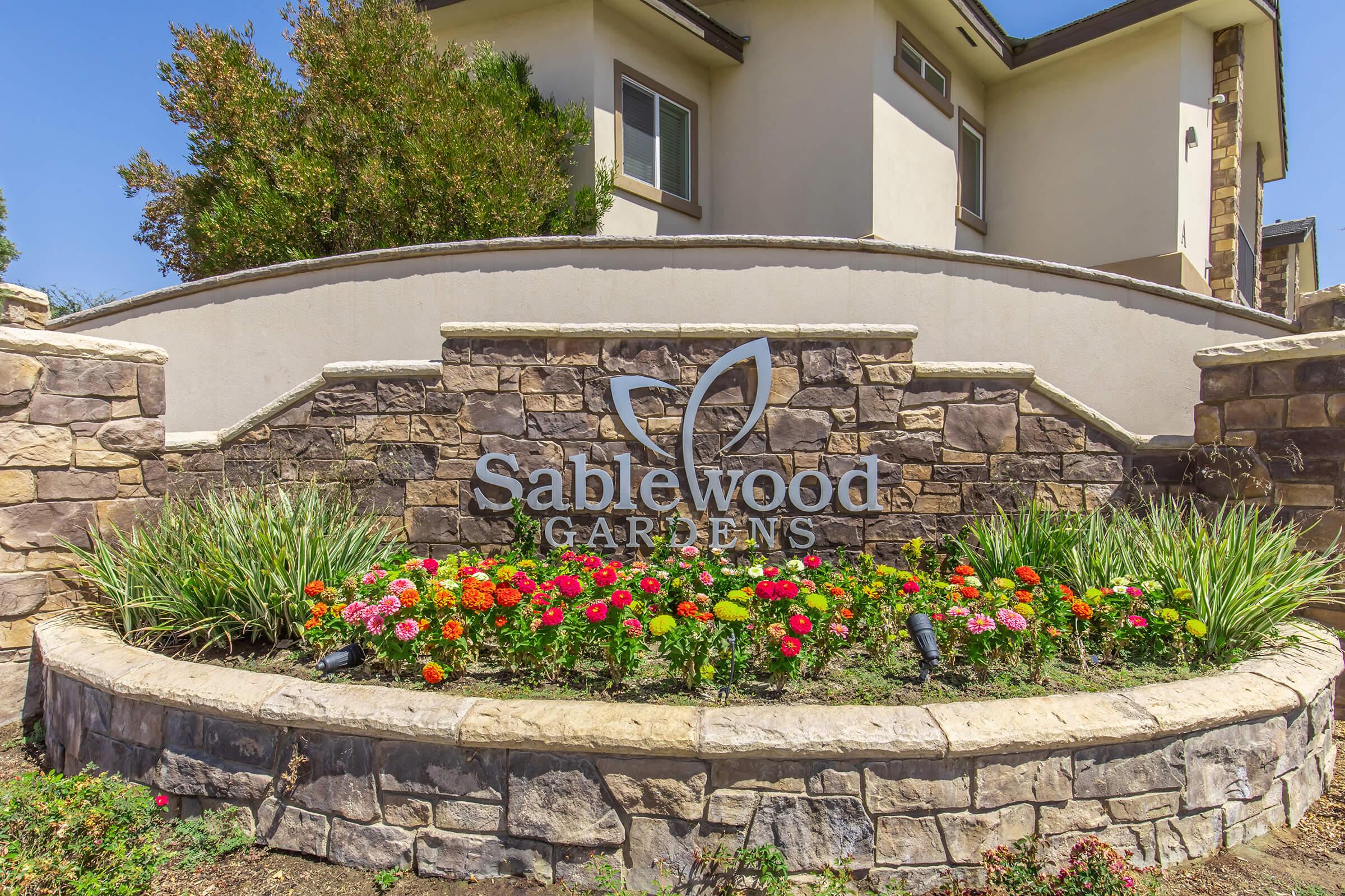 Stone wall with a curved top featuring the logo "Sablewood Gardens" in the center. Below the logo, there is a vibrant flower bed with colorful blooms, surrounded by greenery. A modern building is visible in the background, contributing to a pleasant outdoor atmosphere.