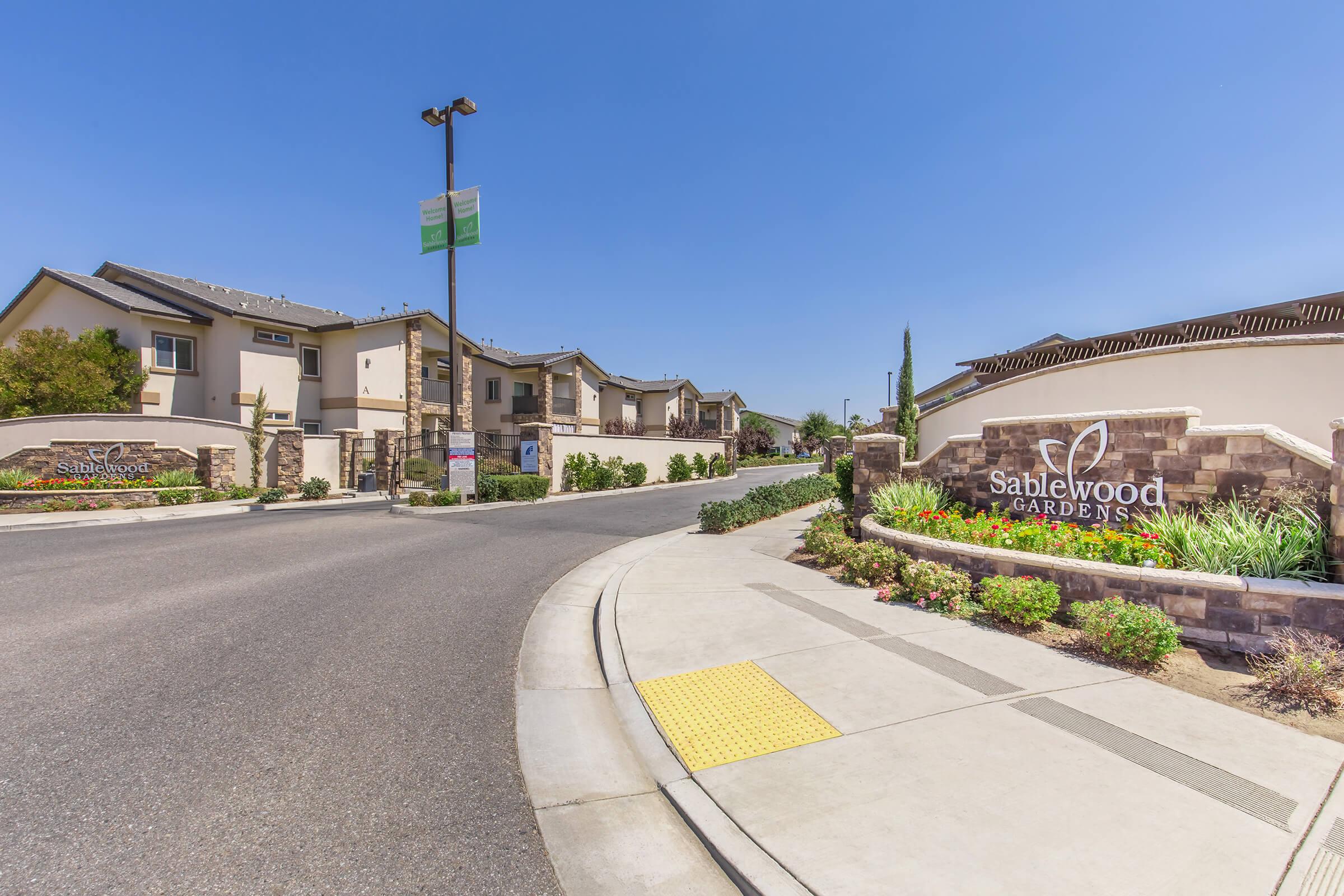 Entrance to Sablewood Gardens, featuring a landscaped area with colorful flowers and shrubs. The sign for Sablewood Gardens is prominently displayed on the right, and residential buildings can be seen in the background under a clear blue sky. The road curves gently, leading into the community.