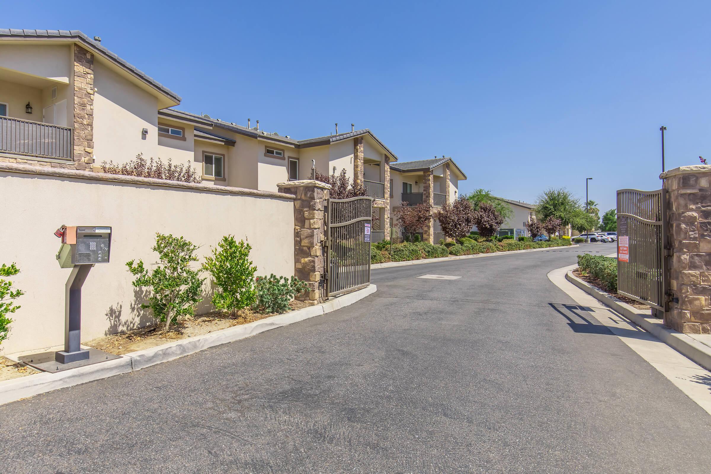 A well-maintained entrance to a residential community featuring a gated entry, a mailbox along the left side, and neatly trimmed bushes. The road curves gently, with houses visible in the background under a clear blue sky.