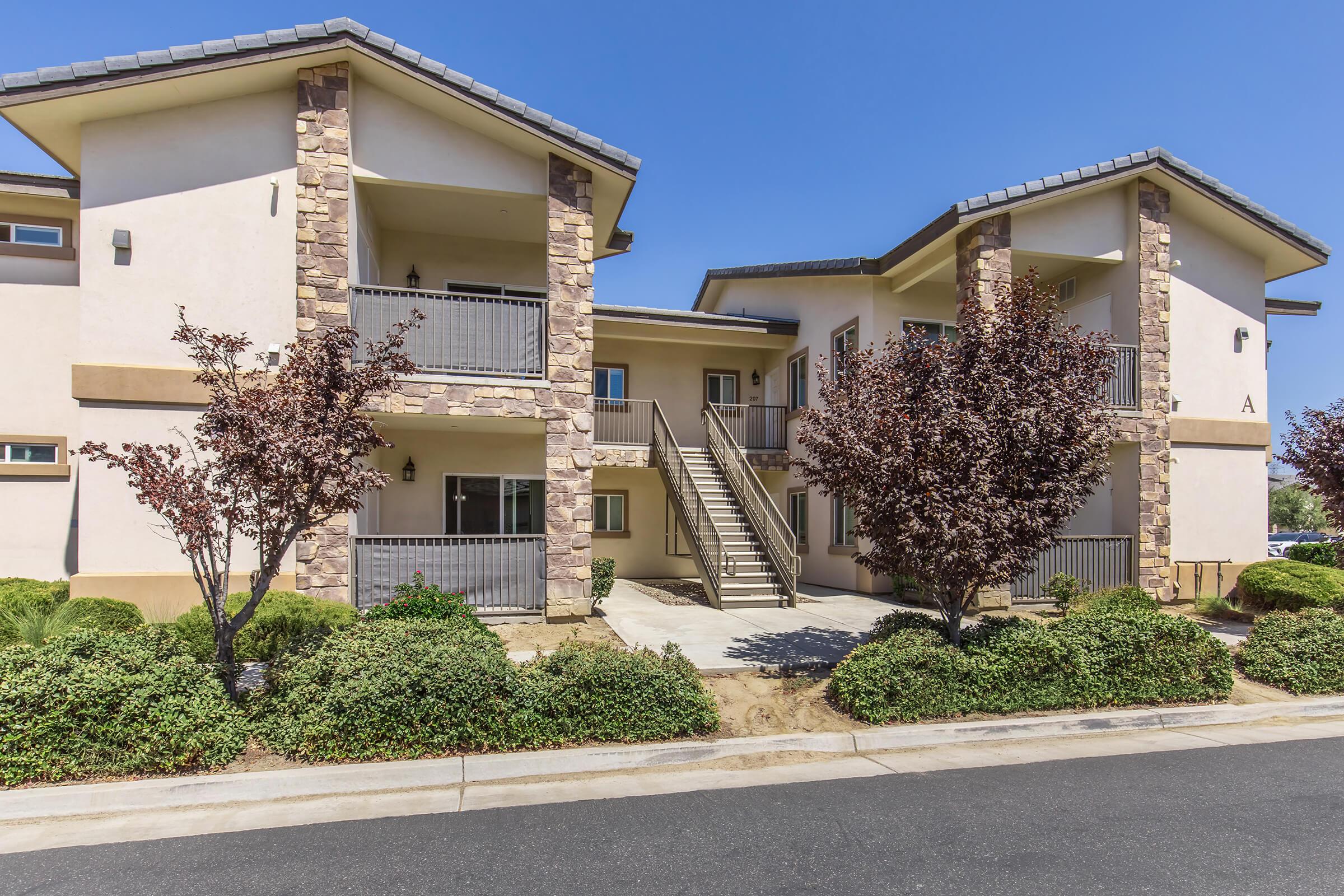 A two-story residential building featuring a balcony on the upper floor, stone accents, and landscaped shrubs. The entrance includes a concrete pathway leading to stairs. The exterior is light-colored, and the sky above is clear and blue.
