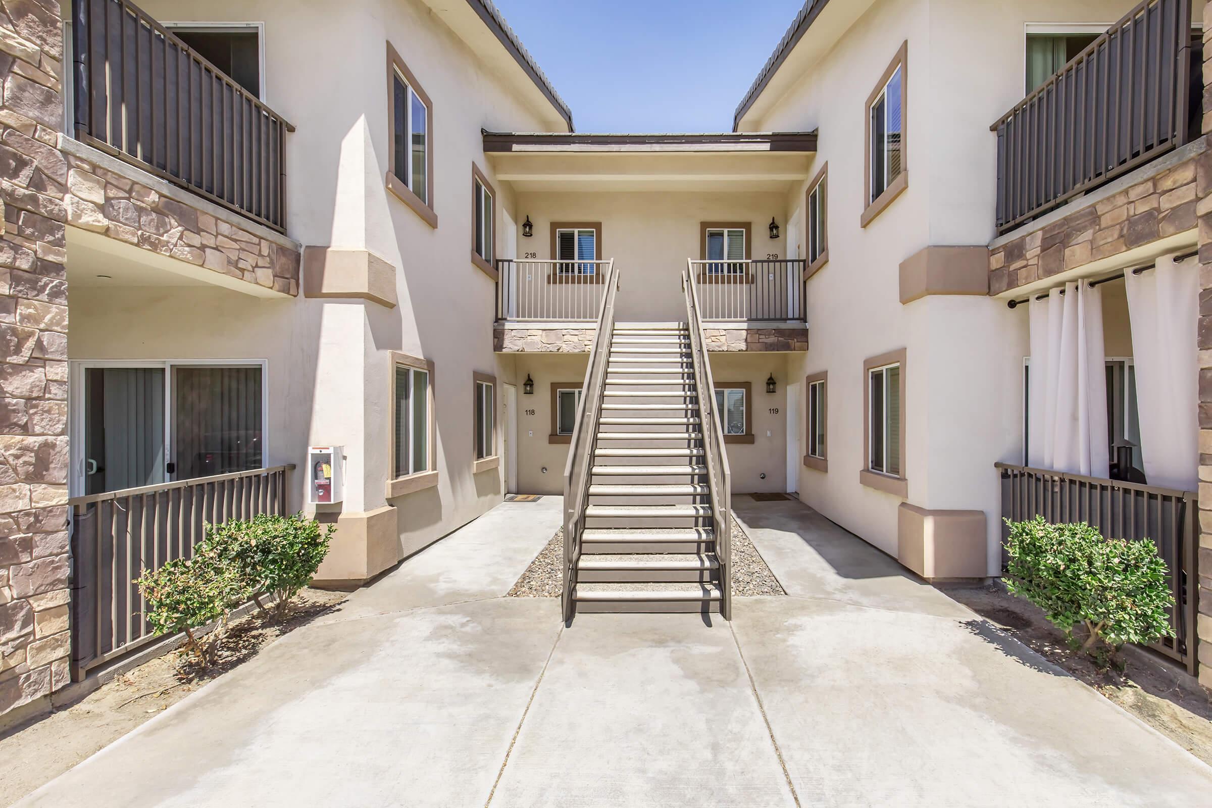 View of a two-story apartment complex featuring a central staircase. The building has a modern design with stone accents and balconies. Green shrubs line the walkway, and large windows are visible on both floors. The sky is clear and blue, creating a bright atmosphere.