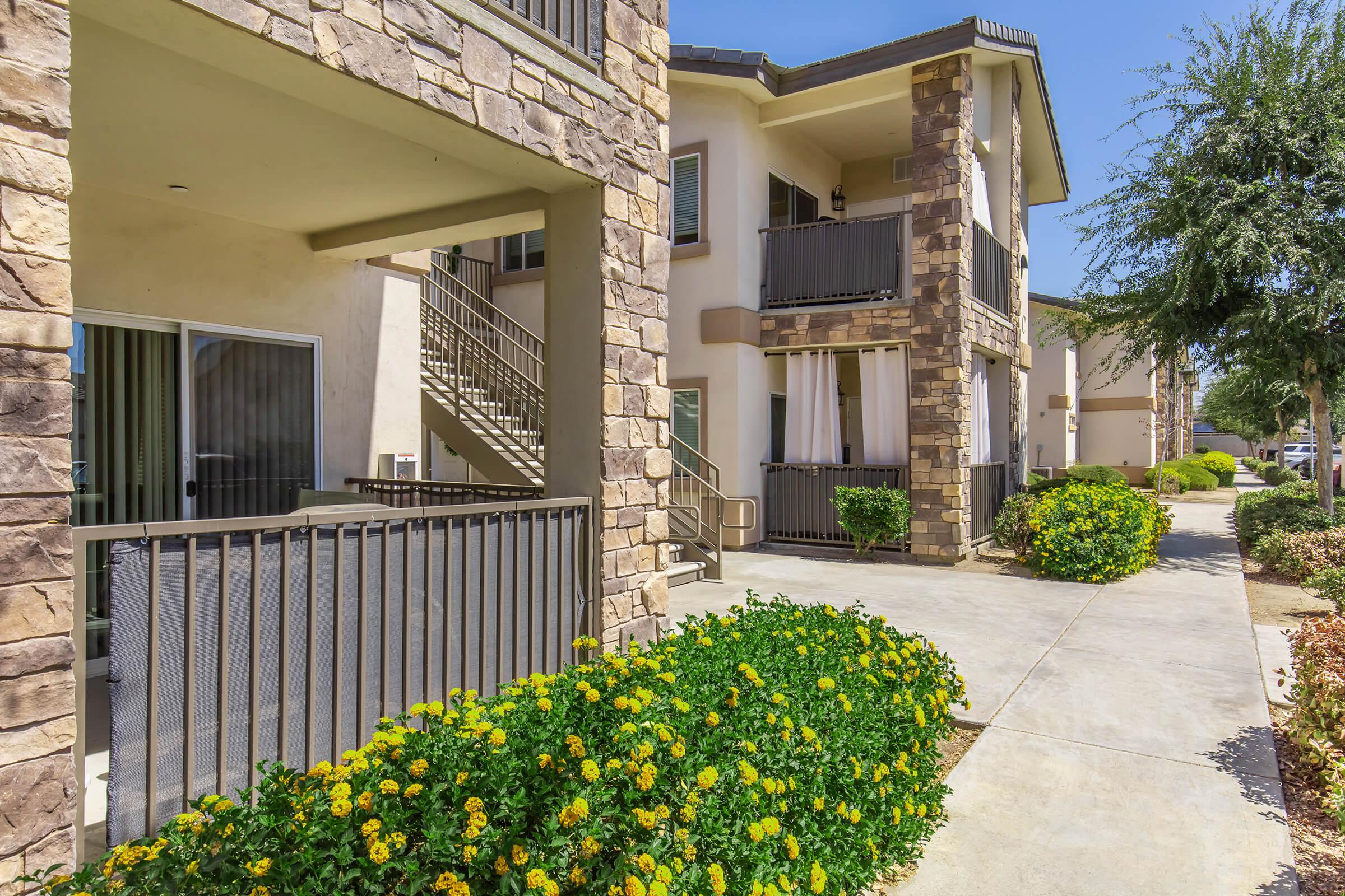 A view of a modern apartment complex featuring two-story buildings with stone accents. The foreground includes a well-maintained pathway flanked by green shrubs and vibrant yellow flower bushes. Balconies are visible on the buildings, along with stairs leading to upper units, all under a clear blue sky.
