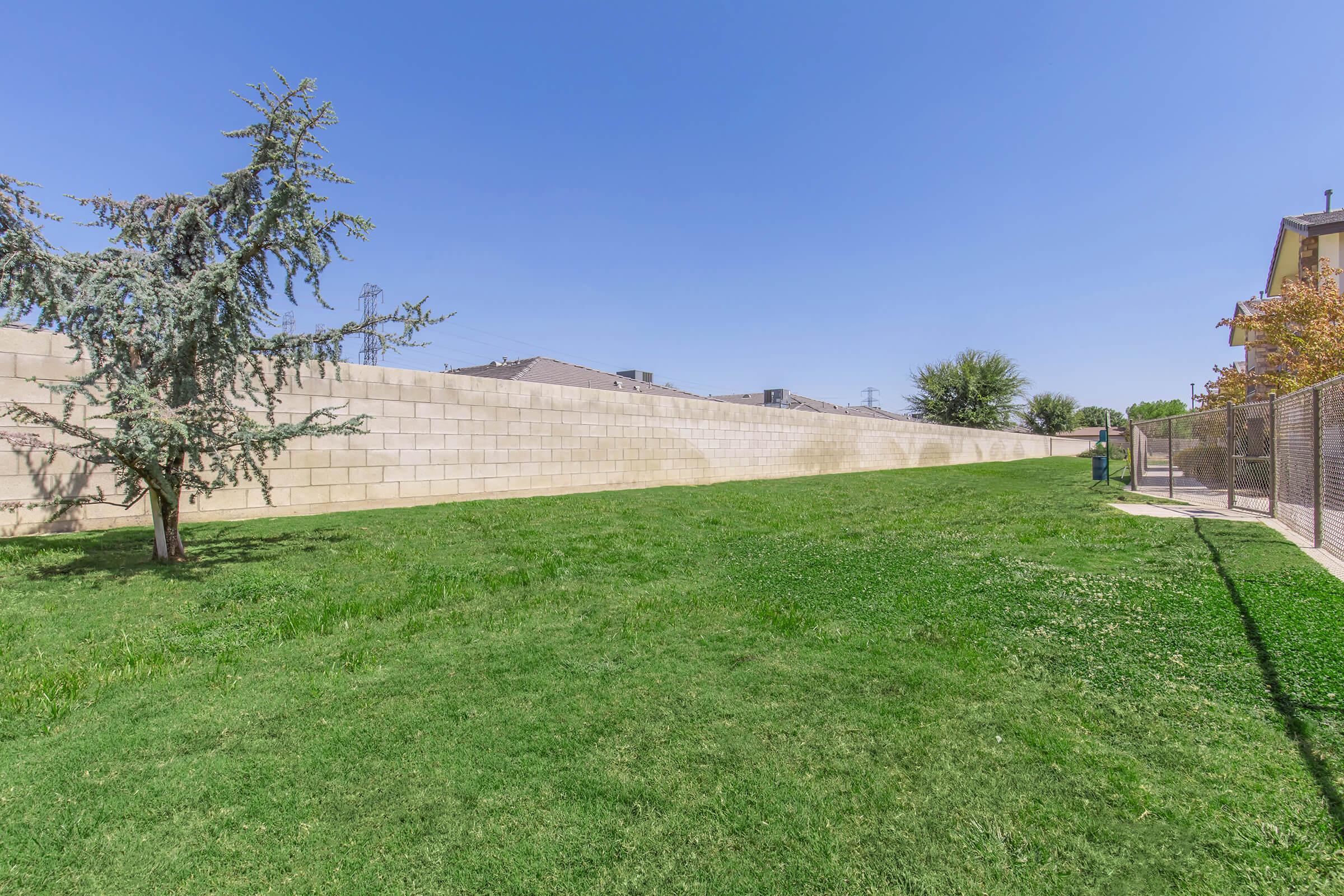 A spacious green lawn with a neatly trimmed grass area, bordered by a tan brick wall on one side. A few trees are visible, and houses can be seen in the background under a clear blue sky. 