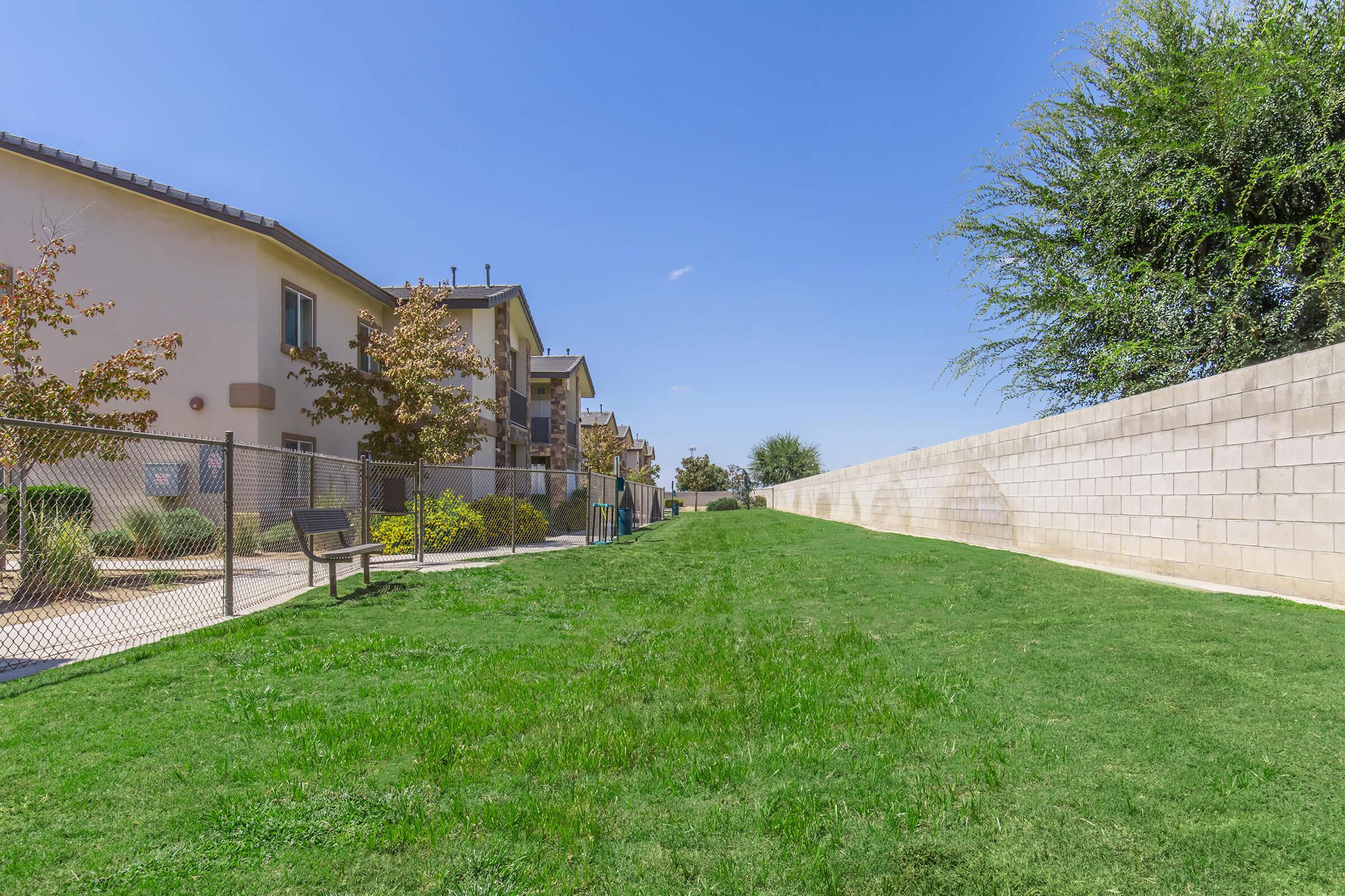 A sunny outdoor scene featuring a grassy pathway between residential buildings and a wall. There are a few benches near the grass, and trees with sparse leaves are visible. The sky is clear blue, suggesting a warm day. The area appears well-maintained and inviting for leisure or walks.