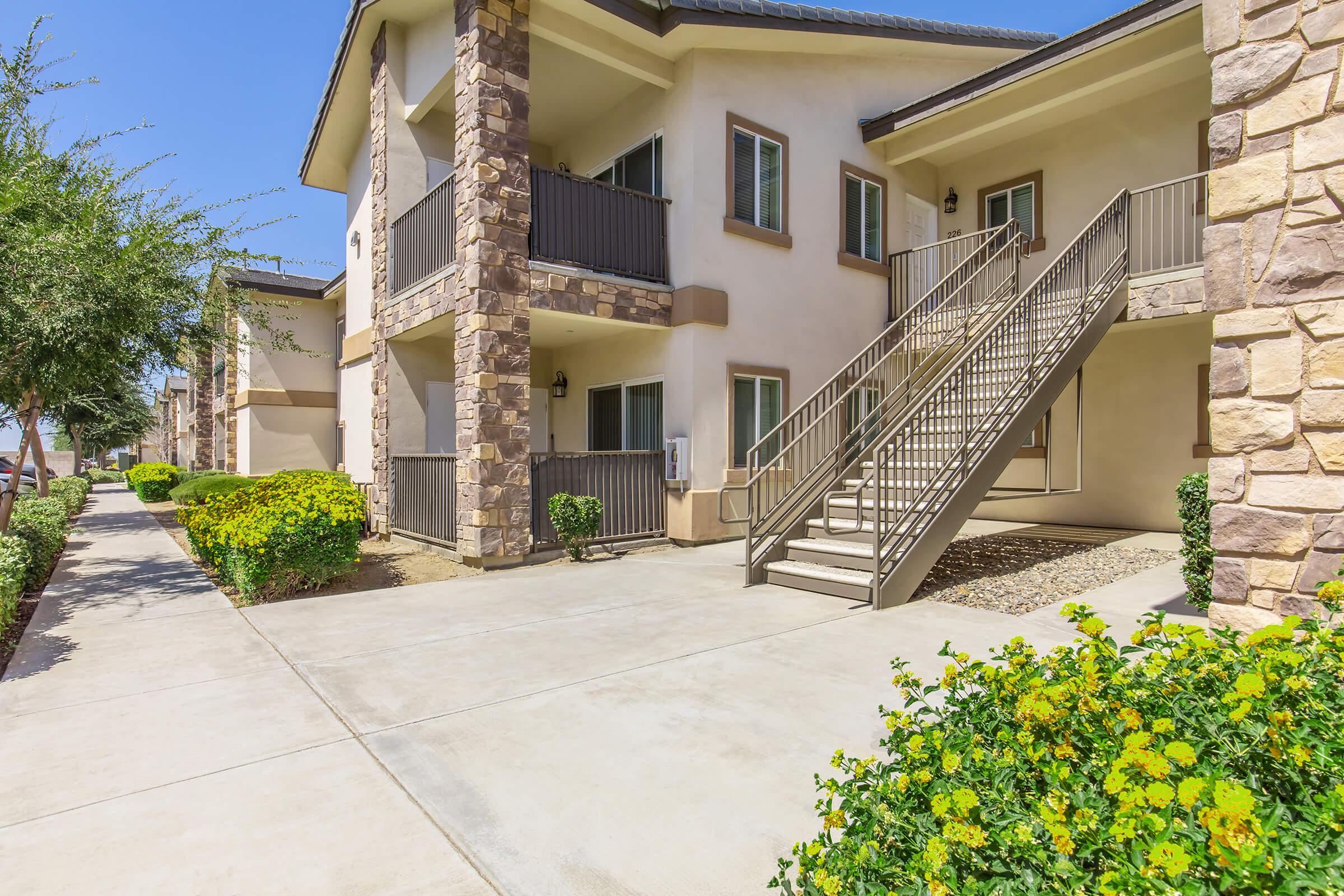 A view of a modern apartment complex featuring multiple two-story buildings with a stone facade. There are visible staircases leading to the second-floor balconies, well-maintained walkways, and landscaped bushes with yellow flowers in the foreground. The sky is clear and sunny.