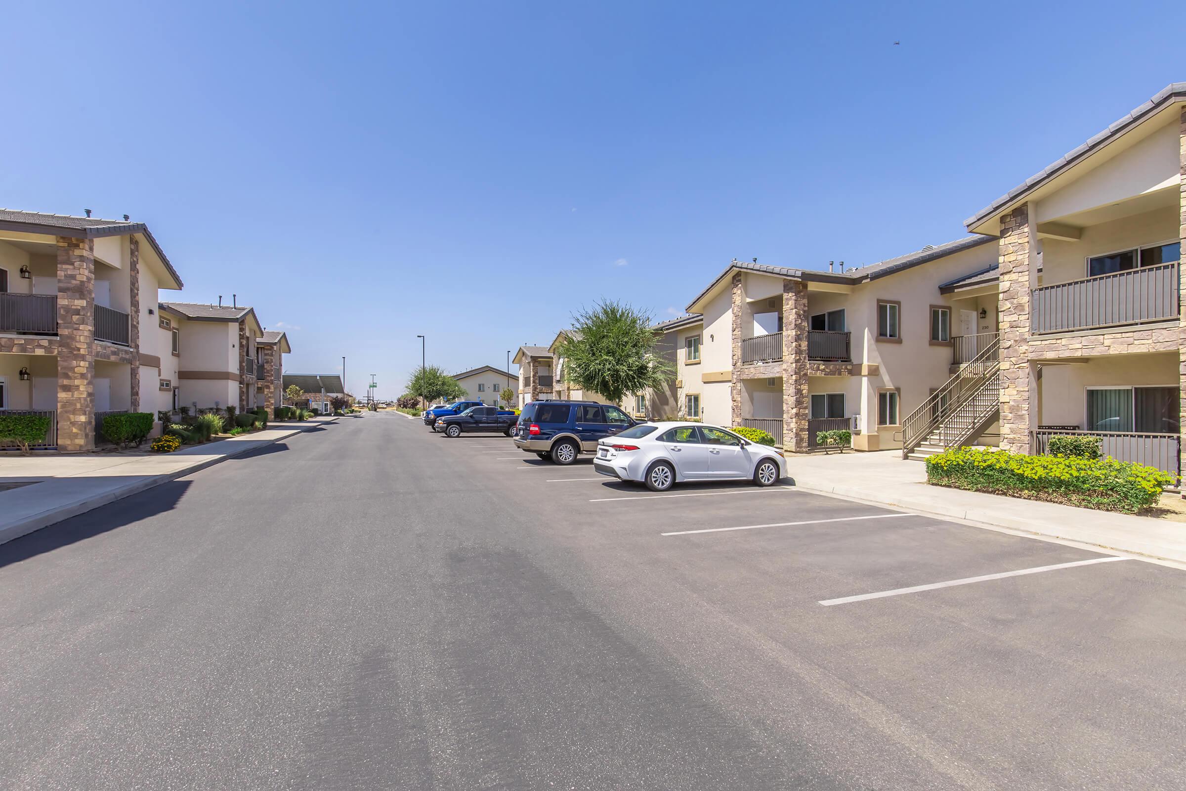 A view of a residential street lined with apartment buildings featuring stone accents. The road is well-paved with parked cars on both sides and lush greenery along the walkways. Bright blue sky overhead enhances the welcoming atmosphere of the neighborhood.