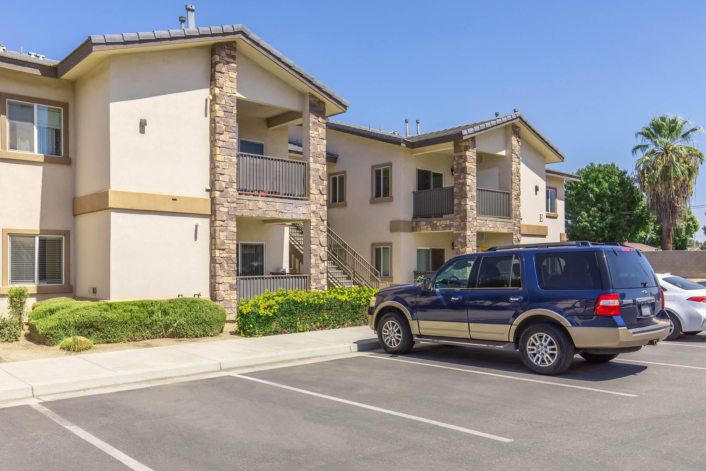 A modern apartment building with a two-story design featuring stone accents. In the foreground, there are parked cars, including a dark SUV. Lush greenery and a palm tree are visible, along with clear blue skies. The scene emphasizes the residential setting.