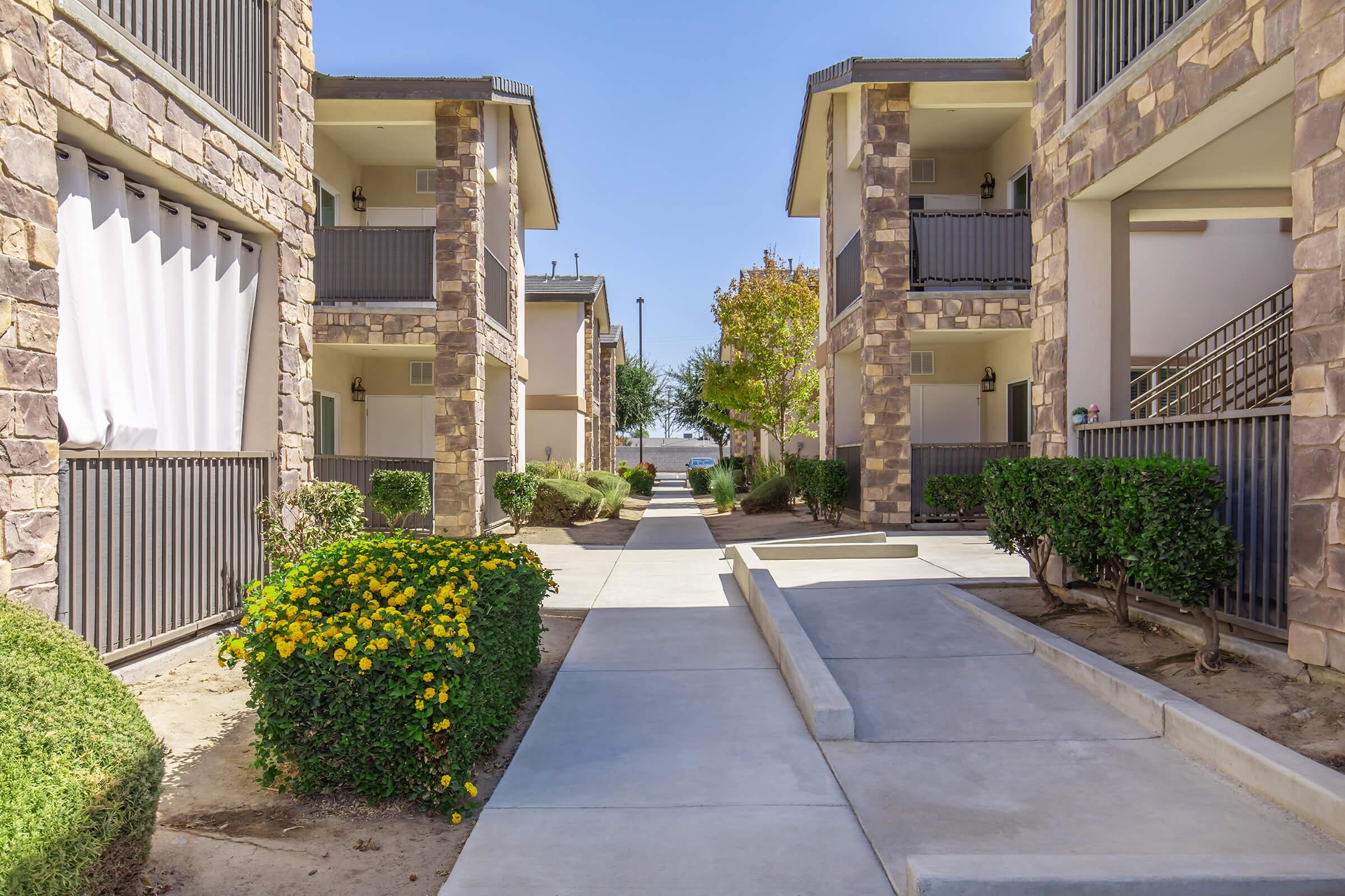 A sunlit pathway between two stone-clad apartment buildings. The walkway is lined with small shrubs and flower bushes, leading to a view of a green area in the background. The sky is clear and blue, highlighting the serene atmosphere of the community space.