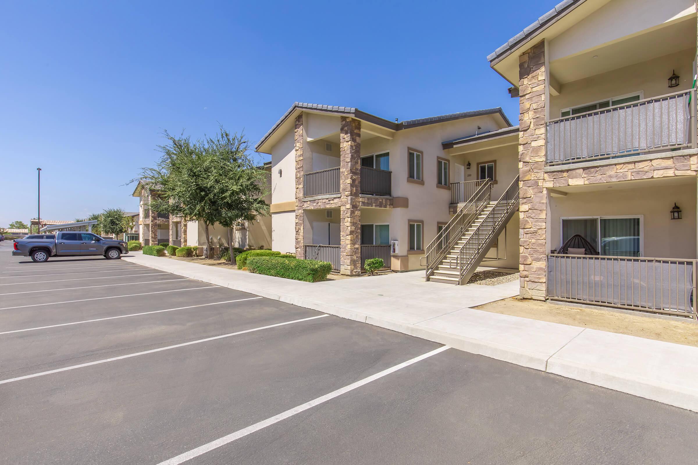 A view of a two-story apartment complex featuring stone accents, a staircase leading to upper units, and a parking area with multiple empty spaces. The building is surrounded by greenery and a clear blue sky.