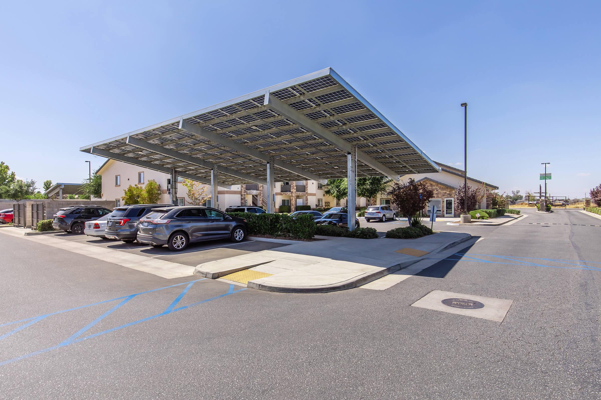 A parking lot featuring solar panel canopies, with several parked vehicles, surrounded by landscaped areas and buildings in the background. The scene is set under a clear blue sky, showcasing modern eco-friendly infrastructure.