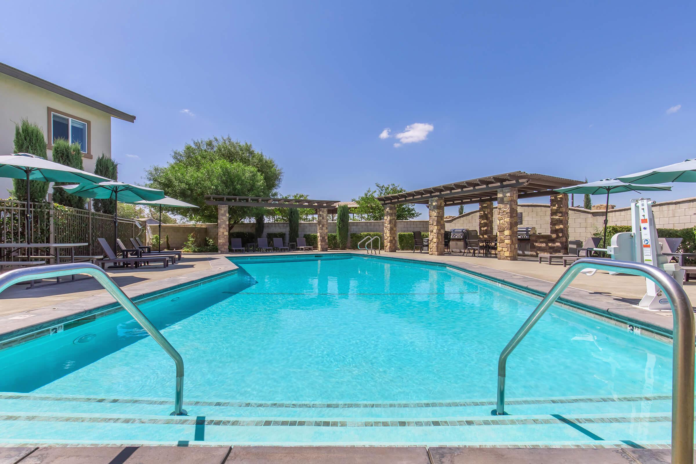 A clear blue swimming pool with metal handrails, surrounded by lounge chairs and umbrellas. In the background, there are shaded areas with stone pillars and green landscaping under a bright blue sky with a few clouds. The scene suggests a relaxing outdoor space for leisure.