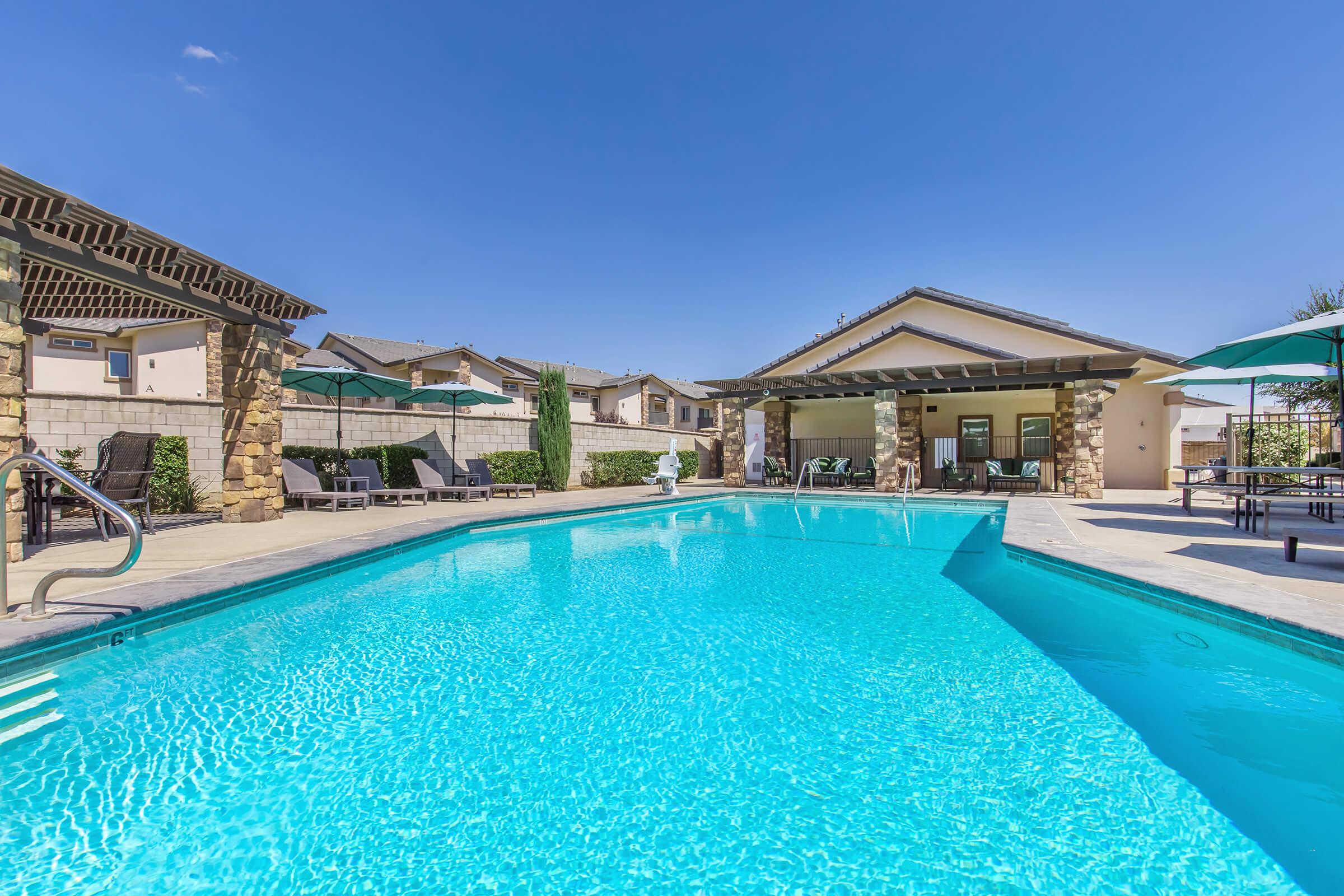 A clear blue swimming pool surrounded by lounge chairs and umbrellas, with a residential building in the background. The sky is sunny and bright, creating a relaxing outdoor atmosphere ideal for leisure activities. Green landscaping is visible alongside the pool area.