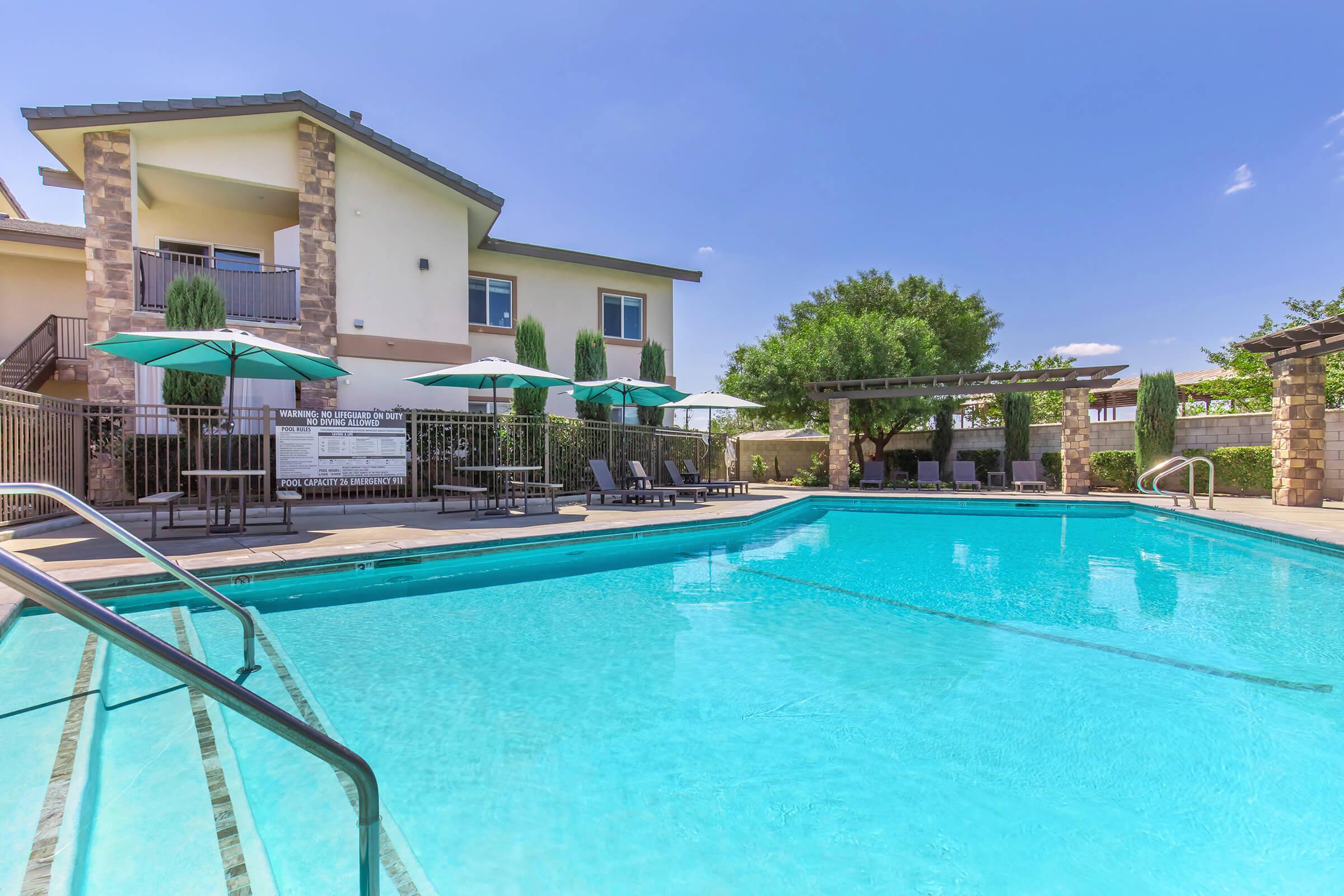 A resort-style swimming pool surrounded by lounge chairs and umbrellas, with a modern building in the background. The pool has steps leading down into the water, and there are green trees and landscaped areas nearby, creating a relaxing outdoor space. The sky is clear and sunny.
