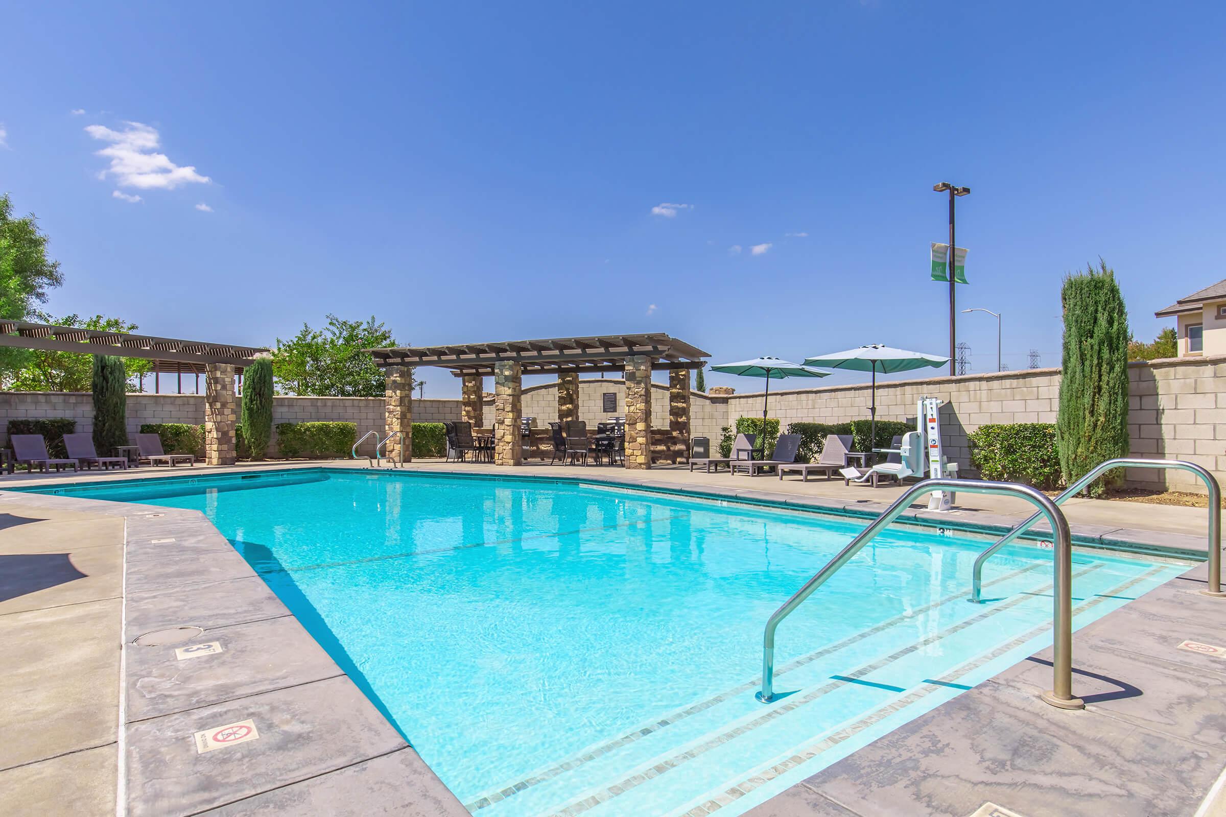 A clear blue swimming pool surrounded by lounge chairs and shaded areas. The pool area features decorative stone structures and green landscaping. Bright blue sky with a few clouds overhead. Ideal for relaxation and leisure activities.