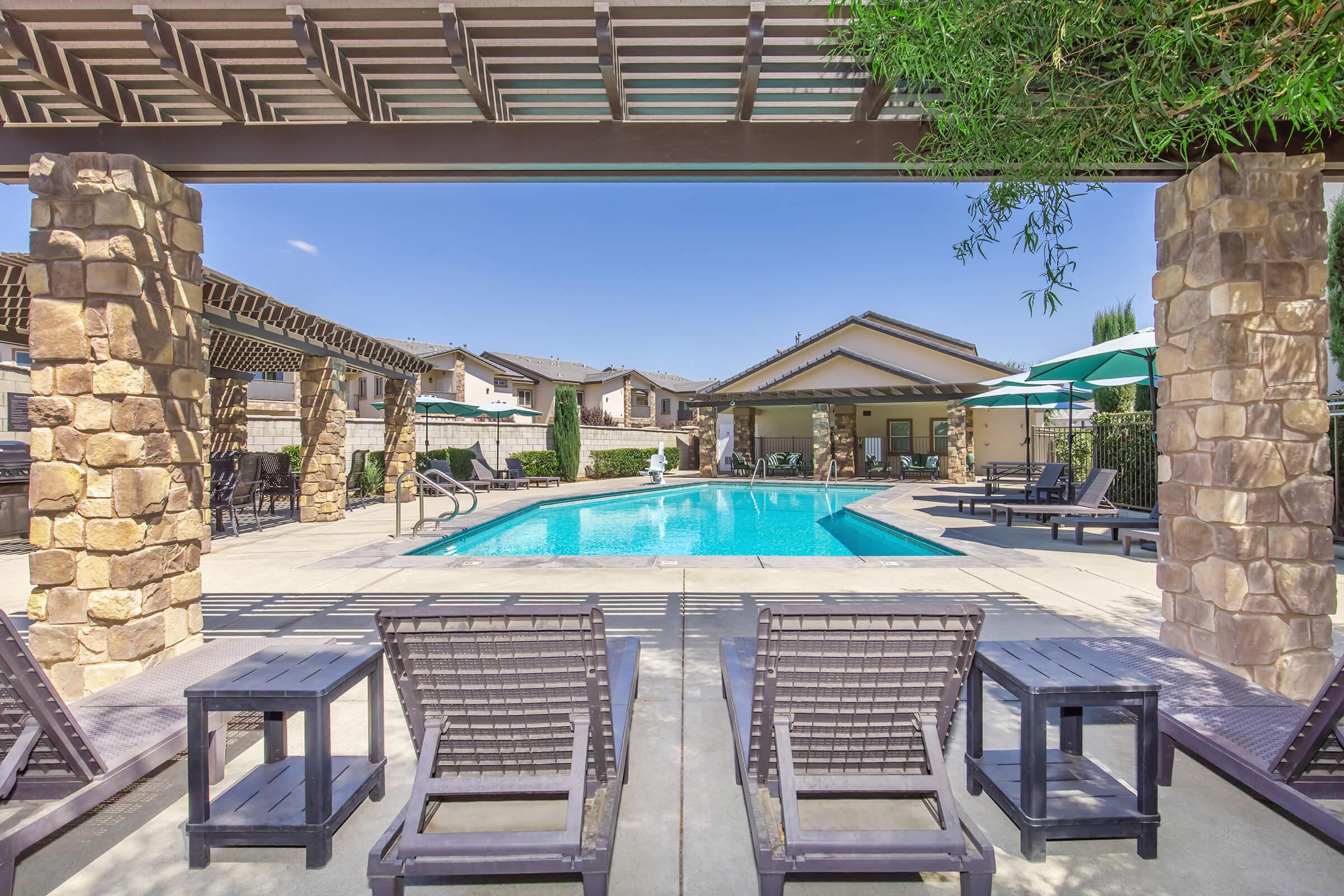 A sunny outdoor pool area featuring a clear blue pool surrounded by lounge chairs and shaded seating. There is a pergola and a nearby building, with umbrellas providing shade, creating a relaxing atmosphere for leisure and recreation.