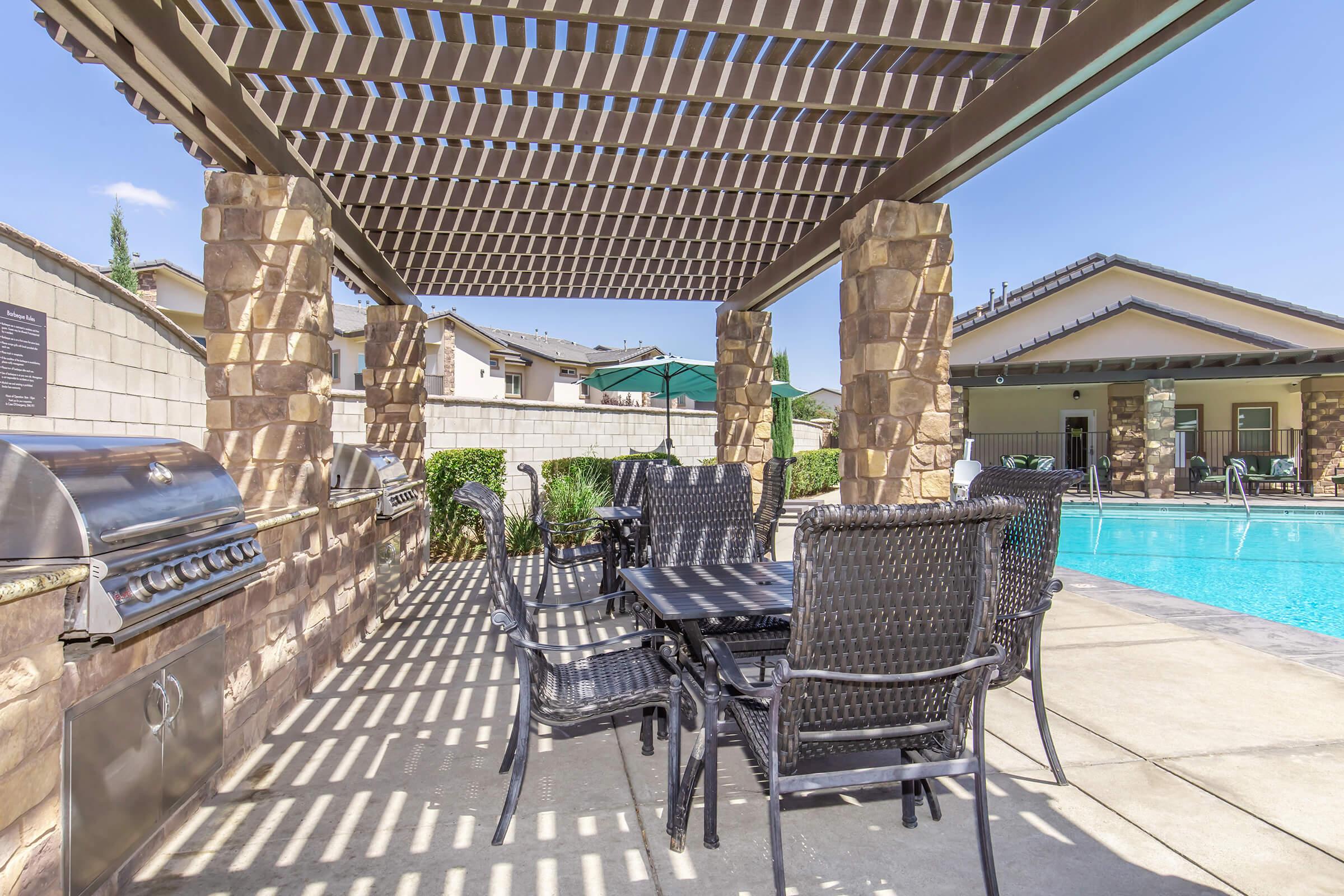 A shaded outdoor area with a stone patio featuring a grill and a dining table surrounded by black wicker chairs. In the background, a swimming pool is visible with clear blue water and a few green umbrellas, along with residential buildings and landscaped greenery.